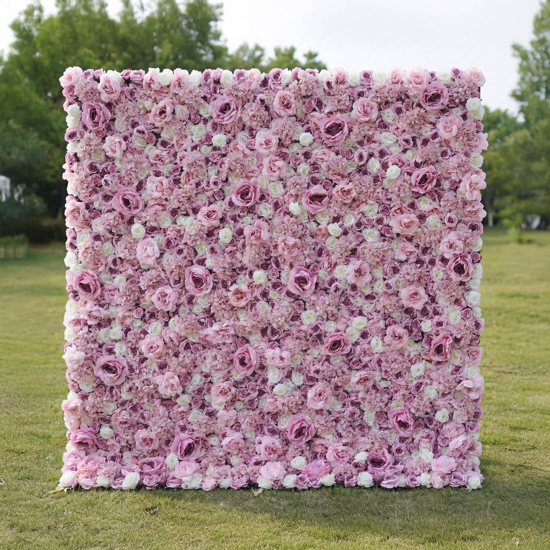 This outdoor floral backdrop rests on a sunlit green lawn, framed by leafy trees. It is a dense, textured wall of artificial blooms in soft blush pink and ivory, featuring layered roses and delicate filler flowers that create a dreamy, romantic surface. The gentle gradient palette and uniform, long-lasting artificial construction make it ideal for garden weddings, proposal setups, or photo booths seeking a feminine, timeless focal point.
