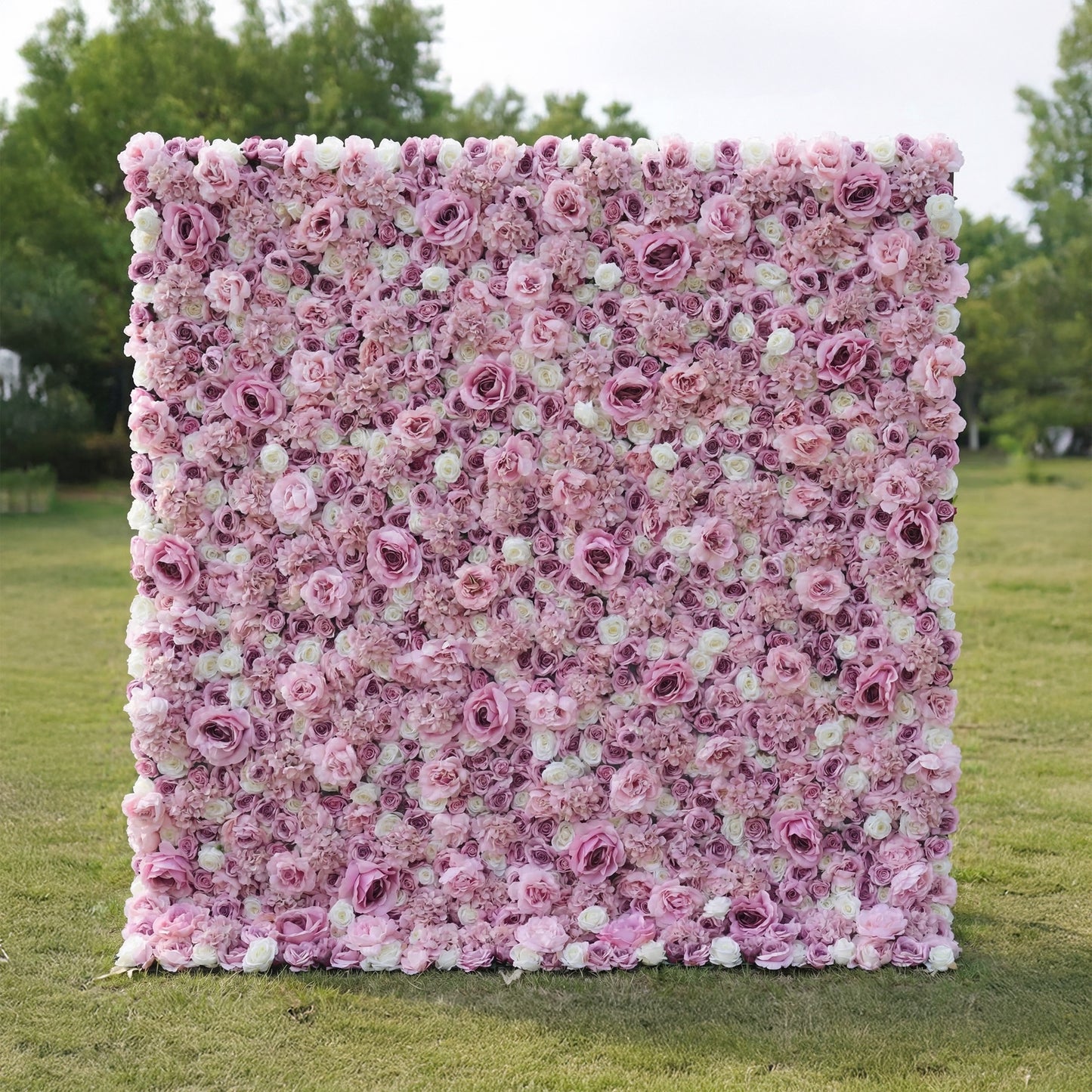 This outdoor floral backdrop rests on a sunlit green lawn, framed by leafy trees. It is a dense, textured wall of artificial blooms in soft blush pink and ivory, featuring layered roses and delicate filler flowers that create a dreamy, romantic surface. The gentle gradient palette and uniform, long-lasting artificial construction make it ideal for garden weddings, proposal setups, or photo booths seeking a feminine, timeless focal point.
