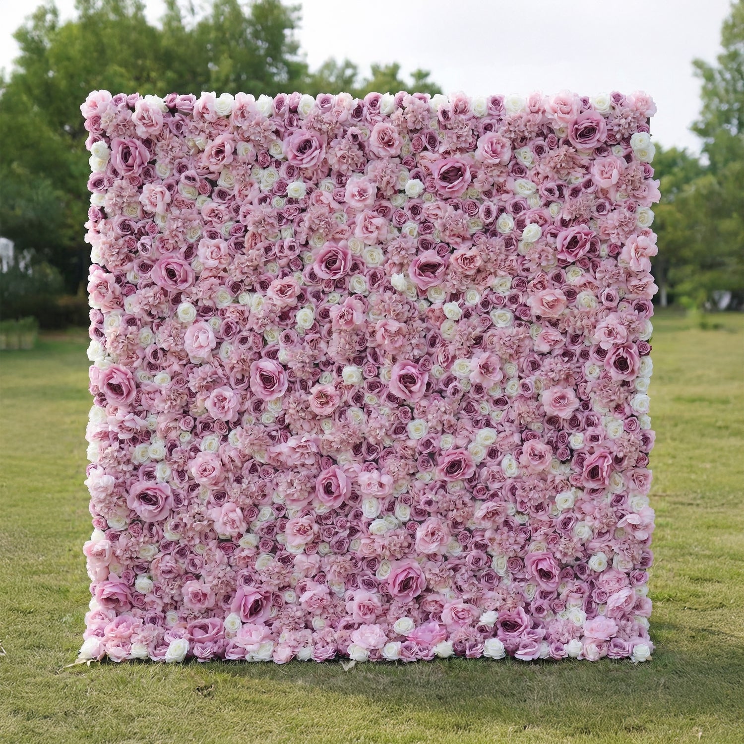 This outdoor floral backdrop rests on a sunlit green lawn, framed by leafy trees. It is a dense, textured wall of artificial blooms in soft blush pink and ivory, featuring layered roses and delicate filler flowers that create a dreamy, romantic surface. The gentle gradient palette and uniform, long-lasting artificial construction make it ideal for garden weddings, proposal setups, or photo booths seeking a feminine, timeless focal point.