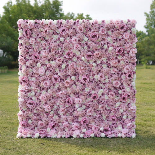 This outdoor floral backdrop rests on a sunlit green lawn, framed by leafy trees. It is a dense, textured wall of artificial blooms in soft blush pink and ivory, featuring layered roses and delicate filler flowers that create a dreamy, romantic surface. The gentle gradient palette and uniform, long-lasting artificial construction make it ideal for garden weddings, proposal setups, or photo booths seeking a feminine, timeless focal point.