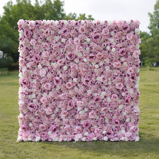 This outdoor floral backdrop rests on a sunlit green lawn, framed by leafy trees. It is a dense, textured wall of artificial blooms in soft blush pink and ivory, featuring layered roses and delicate filler flowers that create a dreamy, romantic surface. The gentle gradient palette and uniform, long-lasting artificial construction make it ideal for garden weddings, proposal setups, or photo booths seeking a feminine, timeless focal point.