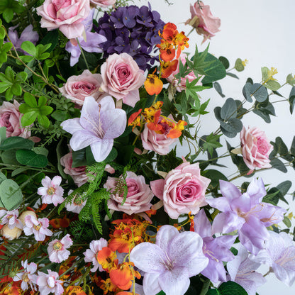 This image showcases an artificial flower arrangement bursting with colors like soft pink, purple, orange, and white. It features roses, hydrangeas, and other delicate blooms. The style is vibrant and romantic, mimicking a lush garden scene. 