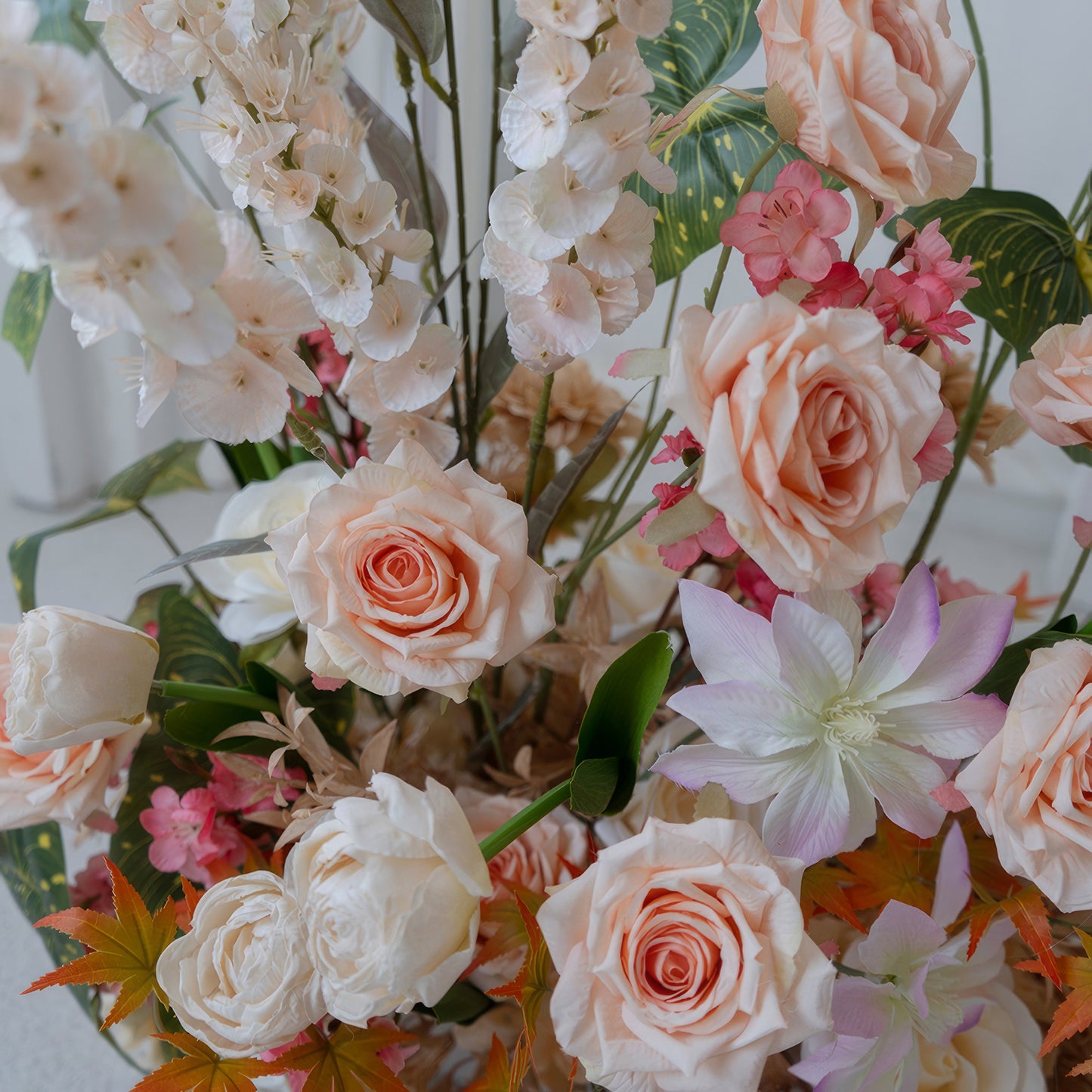 This image showcases an artificial flower arrangement with roses as the prominent type, along with other blossoms. Colors range from soft peach and white to hints of pink and orange, creating a warm and elegant style. 