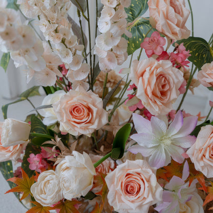 This image showcases an artificial flower arrangement with roses as the prominent type, along with other blossoms. Colors range from soft peach and white to hints of pink and orange, creating a warm and elegant style. 
