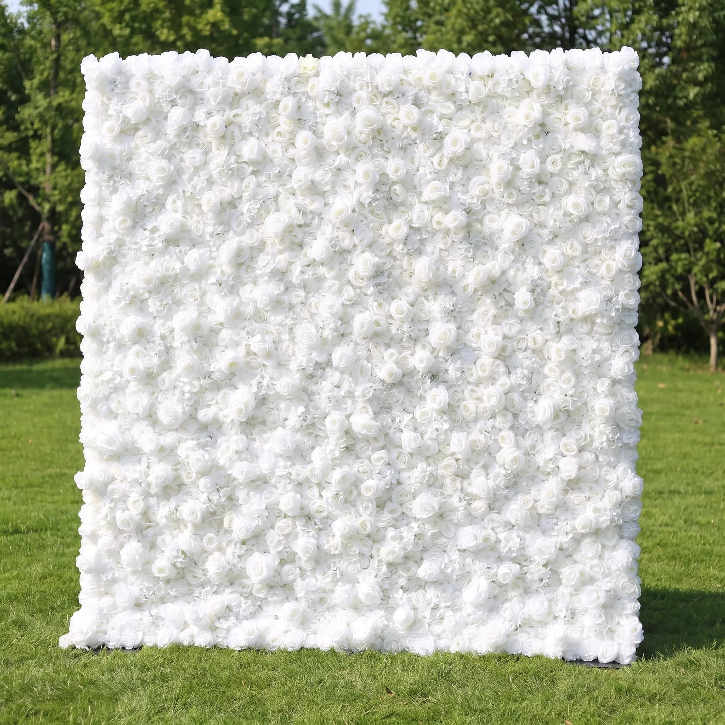 Full dense all-white artificial flower wall covered in layered white roses and delicate airy filler blooms, set on a sunlit green lawn with leafy trees in the background. This reusable, zero-maintenance floral backdrop delivers sleek minimalist luxury, ideal for wedding photo booths, baptism ceremonies, brand boutique displays and neutral clean aesthetic social media content shoots for timeless bright elegant event decor.
