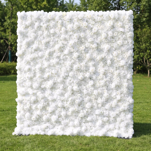 Full dense all-white artificial flower wall covered in layered white roses and delicate airy filler blooms, set on a sunlit green lawn with leafy trees in the background. This reusable, zero-maintenance floral backdrop delivers sleek minimalist luxury, ideal for wedding photo booths, baptism ceremonies, brand boutique displays and neutral clean aesthetic social media content shoots for timeless bright elegant event decor.