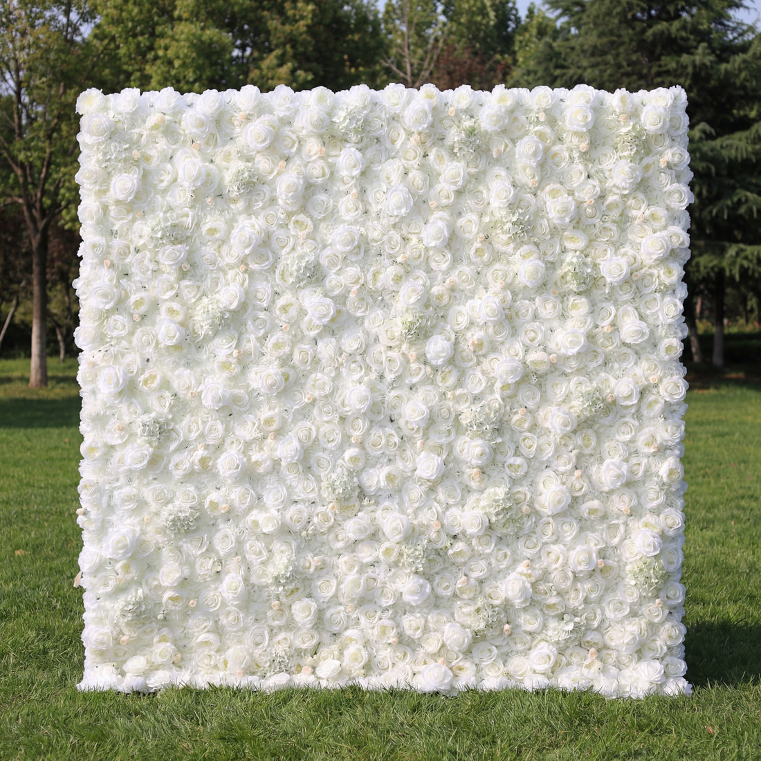 This elegant outdoor floral backdrop features a dense, wall-to-wall arrangement of artificial white roses and delicate filler blooms, creating a lush, velvety texture. Set on a sunlit green lawn and framed by leafy trees, its pure, monochromatic design exudes timeless sophistication and romance, making it a stunning focal point for weddings, engagement photos, or upscale events.