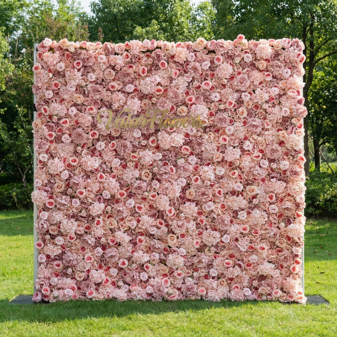 This outdoor floral backdrop stands on a lush green lawn, framed by leafy trees. It features a dense, textured wall of soft blush and ivory roses, creating a dreamy, romantic surface. A polished, cursive gold sign reading “Valar Flowers” anchors the center, contrasting elegantly with the delicate, layered blooms. The soft, feminine palette and timeless design make it perfect for weddings, garden parties, or photo booths seeking an elegant, intimate focal point.