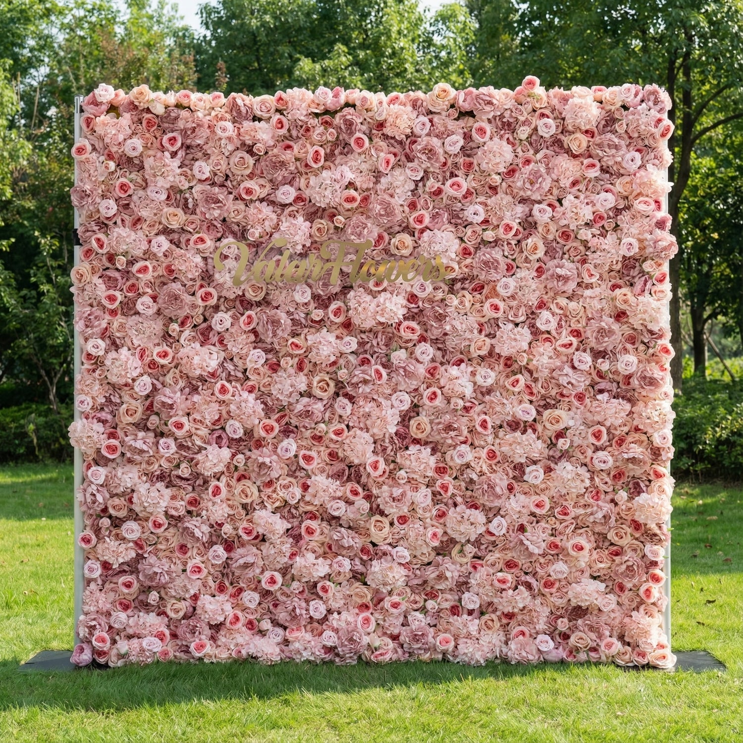This outdoor floral backdrop stands on a lush green lawn, framed by leafy trees. It features a dense, textured wall of soft blush and ivory roses, creating a dreamy, romantic surface. A polished, cursive gold sign reading “Valar Flowers” anchors the center, contrasting elegantly with the delicate, layered blooms. The soft, feminine palette and timeless design make it perfect for weddings, garden parties, or photo booths seeking an elegant, intimate focal point.