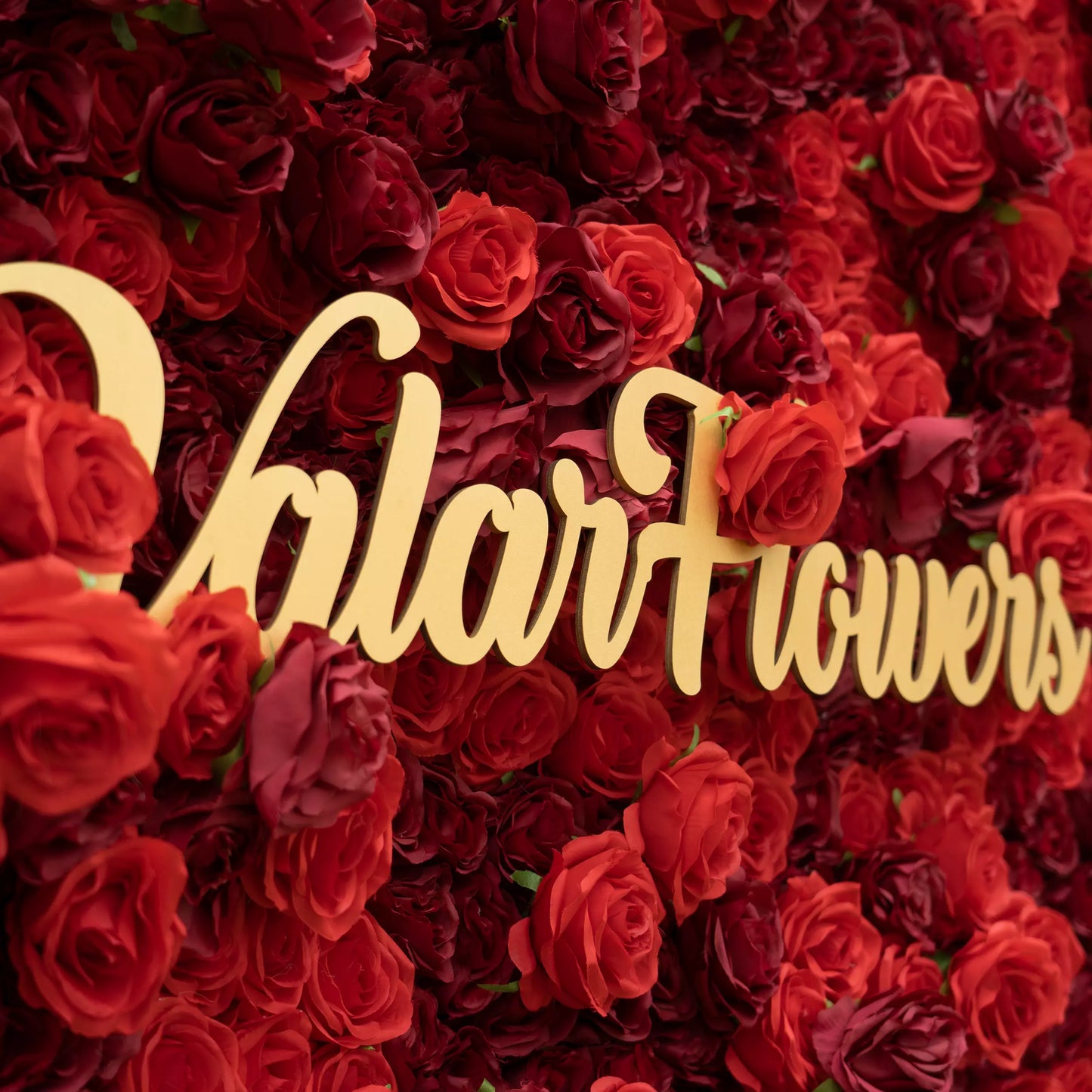 This close-up features a dense backdrop of rich red and burgundy roses, their lush petals creating a romantic, textured display. A polished, cursive "ValarFlowers" sign (metallic gold) anchors the center, contrasting vividly with the deep blooms. The bold, monochromatic palette evokes elegance and warmth, making it ideal for romantic event decor, wedding styling, or premium floral backdrops.