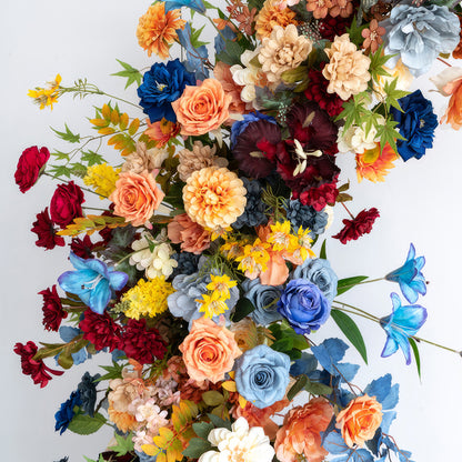 This image showcases a vibrant artificial flower arrangement with a rich color palette of blue, orange, red, yellow, and beige. It features roses, dahlias, and other blooms, creating a lively and festive style. 