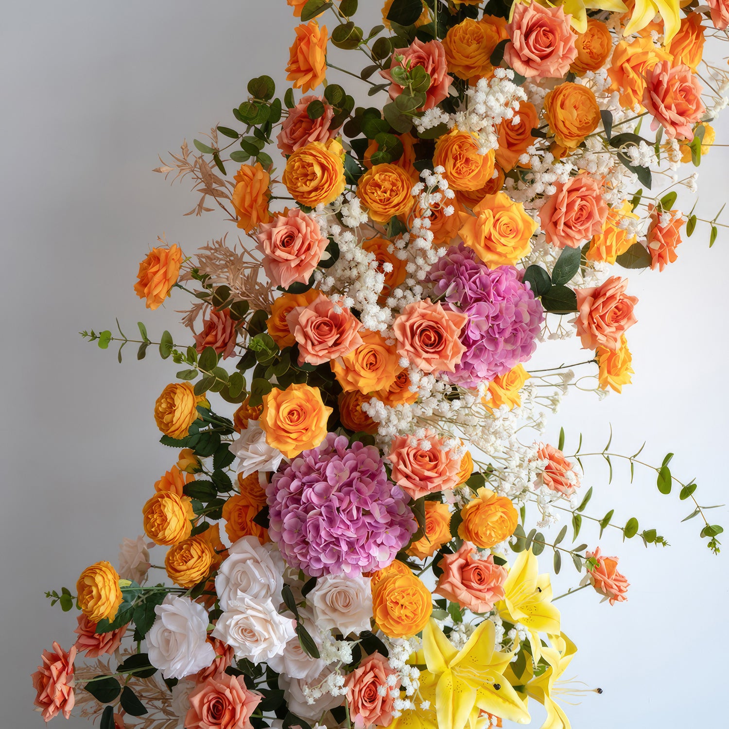 This is a lush floral display with a warm and vivid color scheme. It features an abundance of orange roses, complemented by pops of bright yellow lilies and clusters of purple hydrangeas. White roses and baby&