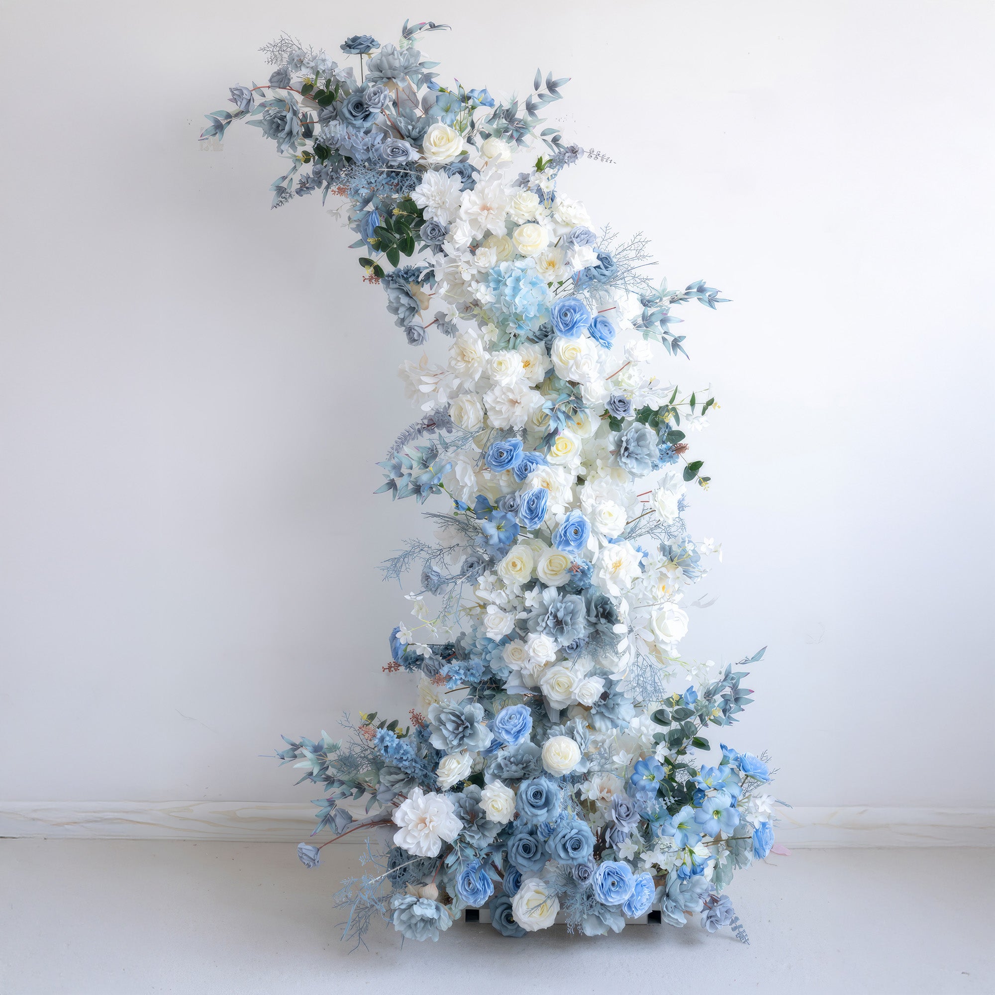 This is a gracefully curved floral installation made of artificial flowers. It combines pure white with various tones of blue, including light and deep blues. Featuring roses and other delicate blooms, along with leafy greens, it conveys a calm, elegant, and romantic style. 