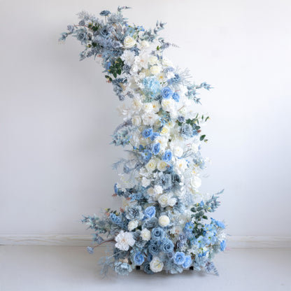 This is a gracefully curved floral installation made of artificial flowers. It combines pure white with various tones of blue, including light and deep blues. Featuring roses and other delicate blooms, along with leafy greens, it conveys a calm, elegant, and romantic style. 