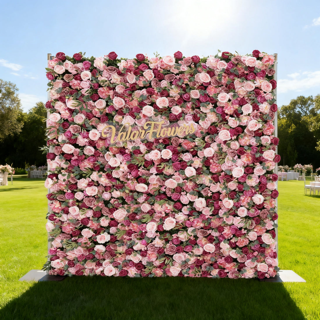 This image features a large, square floral wall composed of roses in various shades of pink and purple. In the center, golden text reads "Valar Flowers". Set outdoors on a lush green lawn under a bright blue sky with scattered clouds, it creates an elegant and romantic atmosphere, perfect for events or decorative displays.