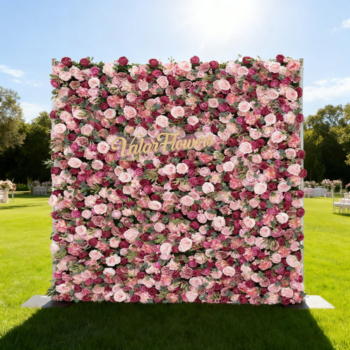 This image features a large, square floral wall composed of roses in various shades of pink and purple. In the center, golden text reads "Valar Flowers". Set outdoors on a lush green lawn under a bright blue sky with scattered clouds, it creates an elegant and romantic atmosphere, perfect for events or decorative displays.