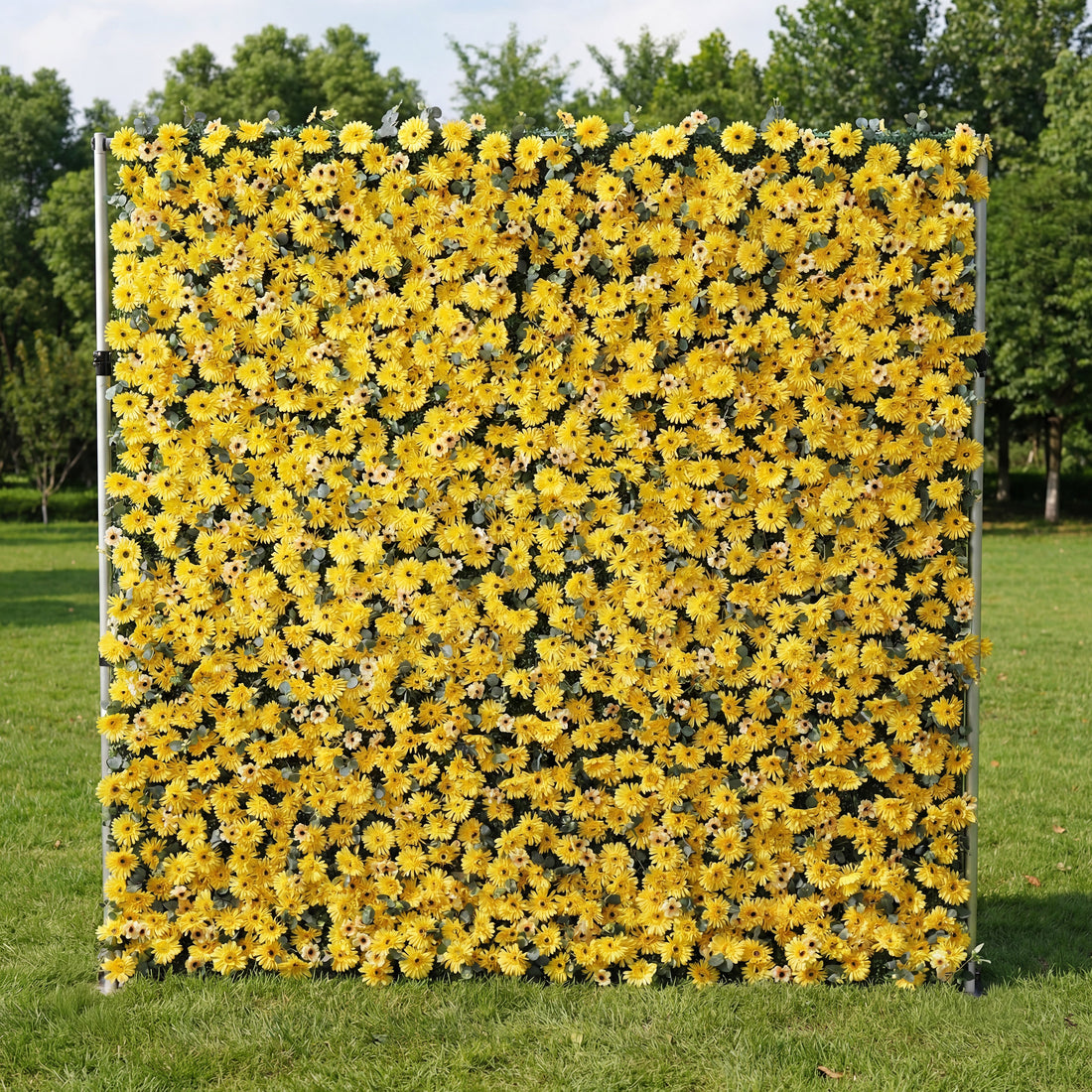 This vibrant outdoor backdrop features a dense, wall-to-wall arrangement of artificial yellow daisies with dark centers, creating a cheerful, sun-drenched texture. Standing on a lush green lawn and framed by leafy trees, its bold, monochromatic design exudes energy and warmth, making it a lively focal point for summer events, photoshoots, or garden parties seeking a bright, joyful aesthetic.