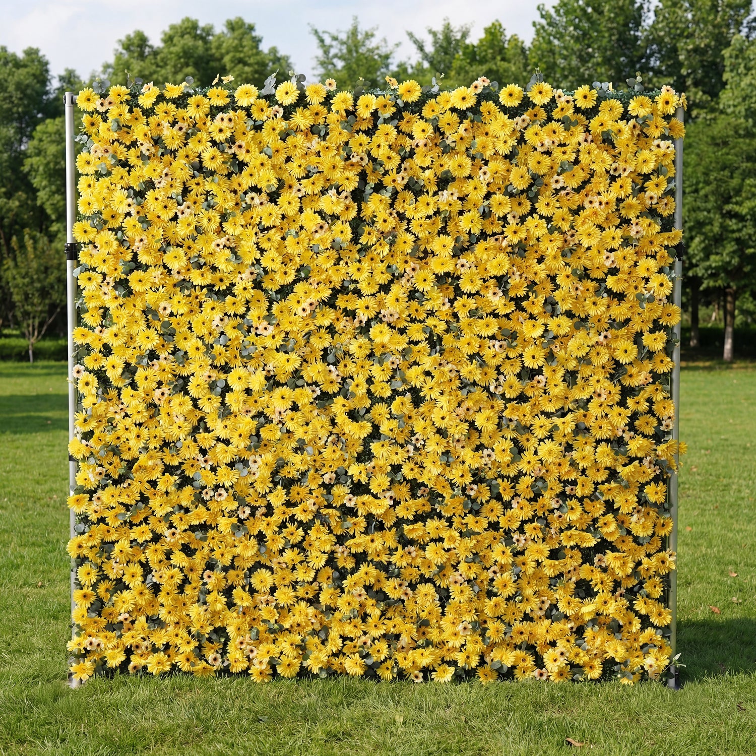 This vibrant outdoor backdrop features a dense, wall-to-wall arrangement of artificial yellow daisies with dark centers, creating a cheerful, sun-drenched texture. Standing on a lush green lawn and framed by leafy trees, its bold, monochromatic design exudes energy and warmth, making it a lively focal point for summer events, photoshoots, or garden parties seeking a bright, joyful aesthetic.