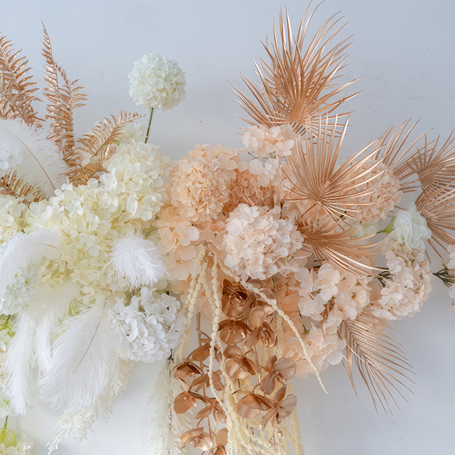 The image features an artificial flower arrangement with hydrangeas, palm fronds, and feathers. Colors include white, peach, and metallic gold - brown. The style is luxurious and modern, ideal for event decoration. 