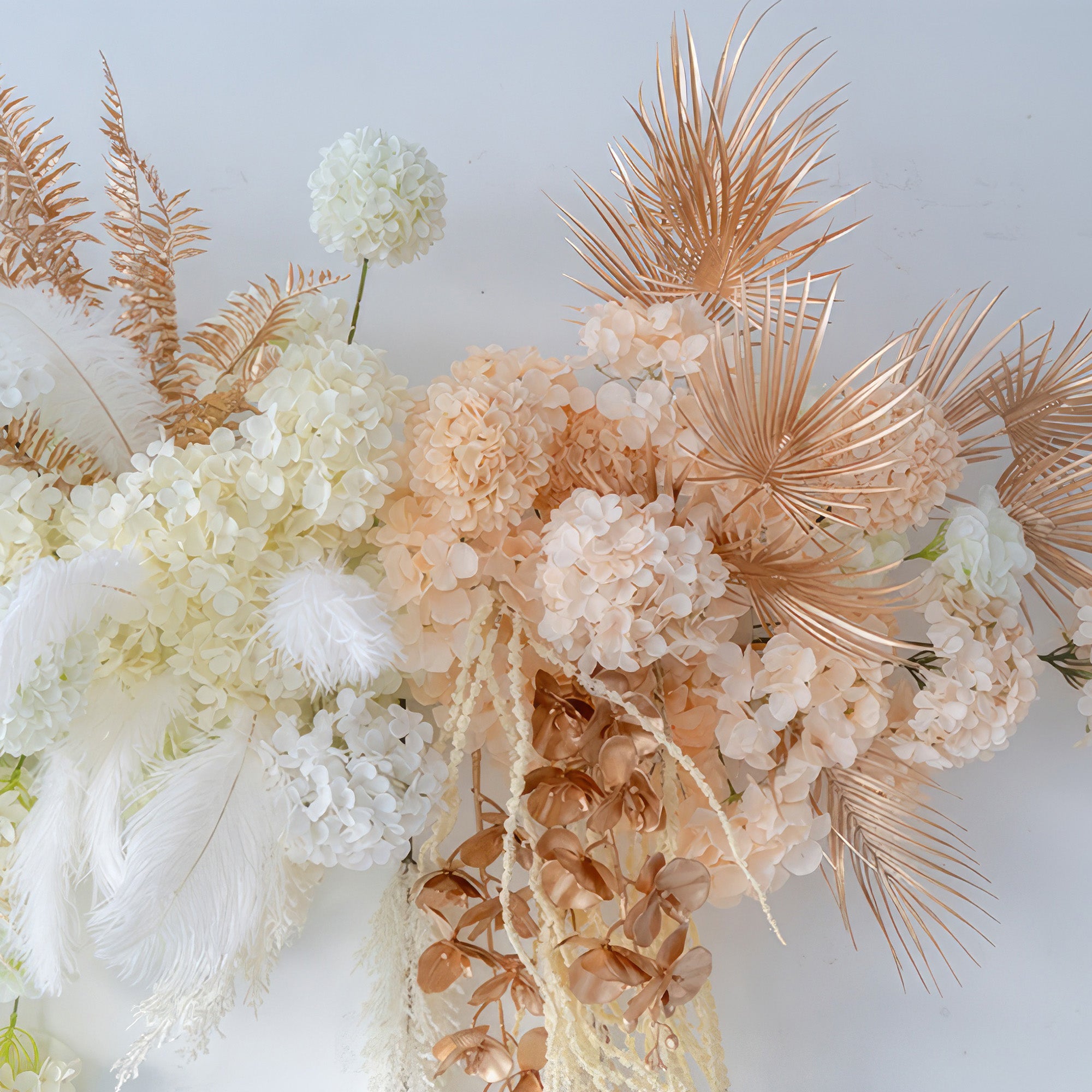 The image features an artificial flower arrangement with hydrangeas, palm fronds, and feathers. Colors include white, peach, and metallic gold - brown. The style is luxurious and modern, ideal for event decoration. 