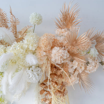 The image features an artificial flower arrangement with hydrangeas, palm fronds, and feathers. Colors include white, peach, and metallic gold - brown. The style is luxurious and modern, ideal for event decoration. 