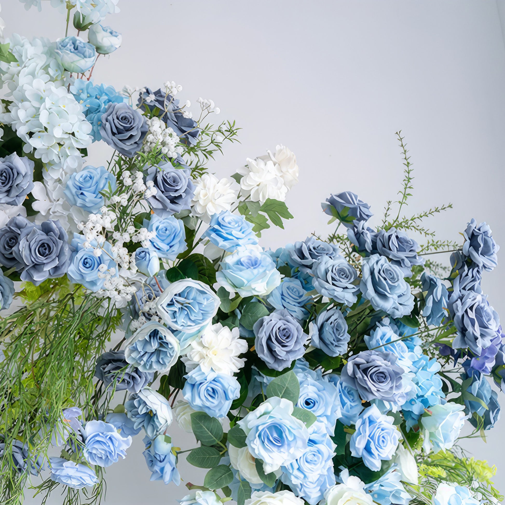 A close-up of a lush floral arrangement from Valar Flowers, featuring a harmonious blend of deep navy, sky blue, and creamy white blooms—including roses, hydrangeas, and delicate baby's breath—intertwined with silvery-green foliage. The layered, textured petals create a sophisticated, cool-toned aesthetic, ideal for wedding decor, centerpieces, or special event accents.