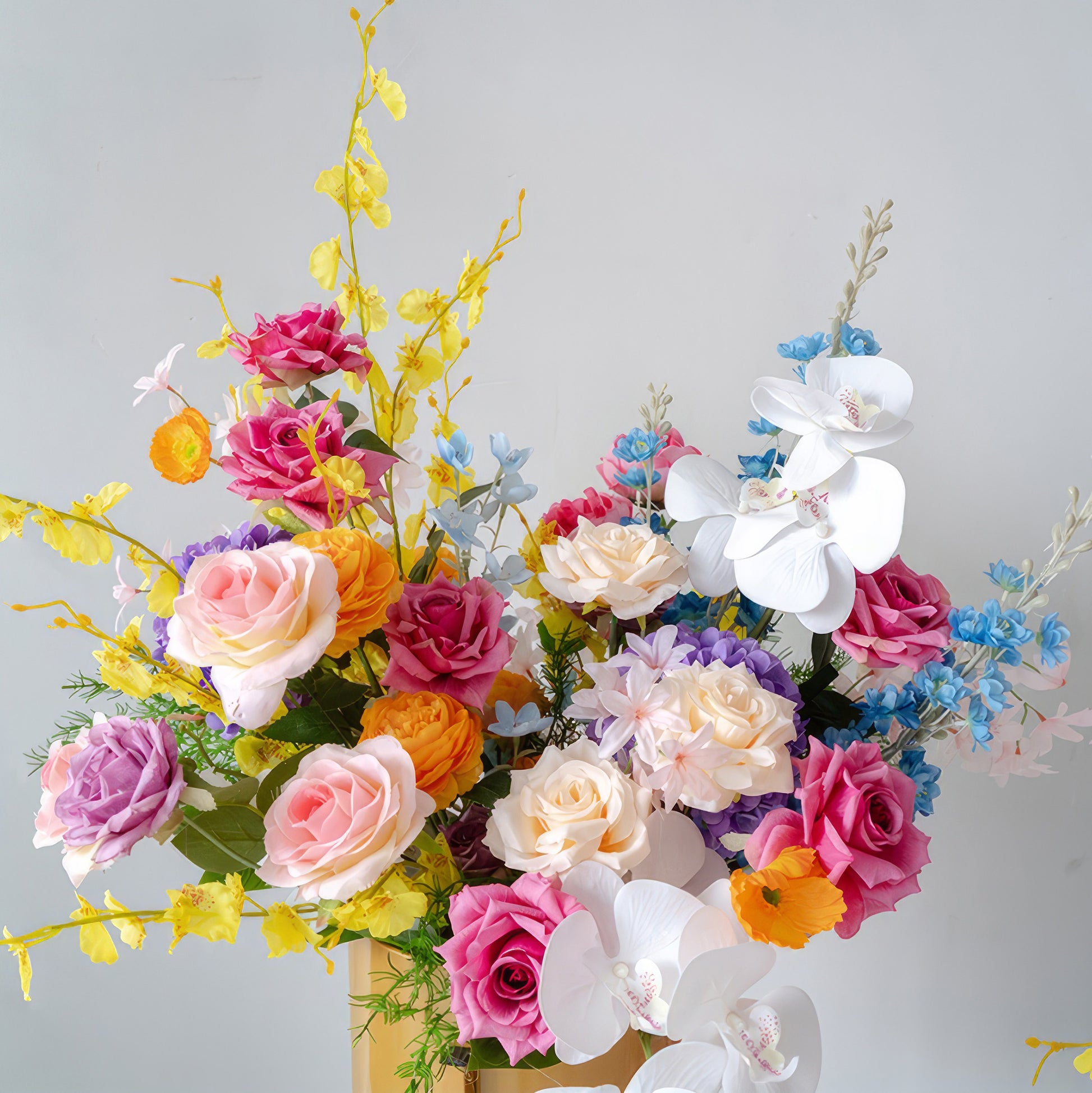 This vibrant floral arrangement features a rich mix of artificial flowers in a plethora of colors. There are roses in shades from soft pastels like pink and lavender to bold tones such as hot pink and orange. White orchids and small blue blossoms add contrast. With green foliage, it exudes a lively, celebratory style. 
