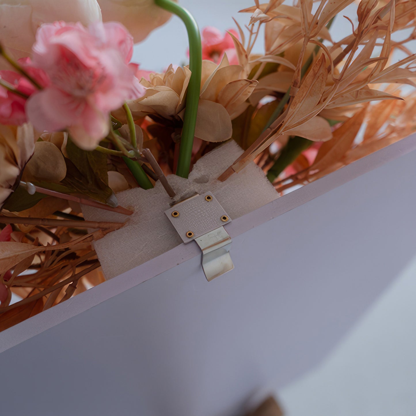 The image displays a close - up of an artificial flower arrangement attached to a board. There's a metal clip with holes, secured by a fabric strip, holding flower stems firmly in place against the board. 