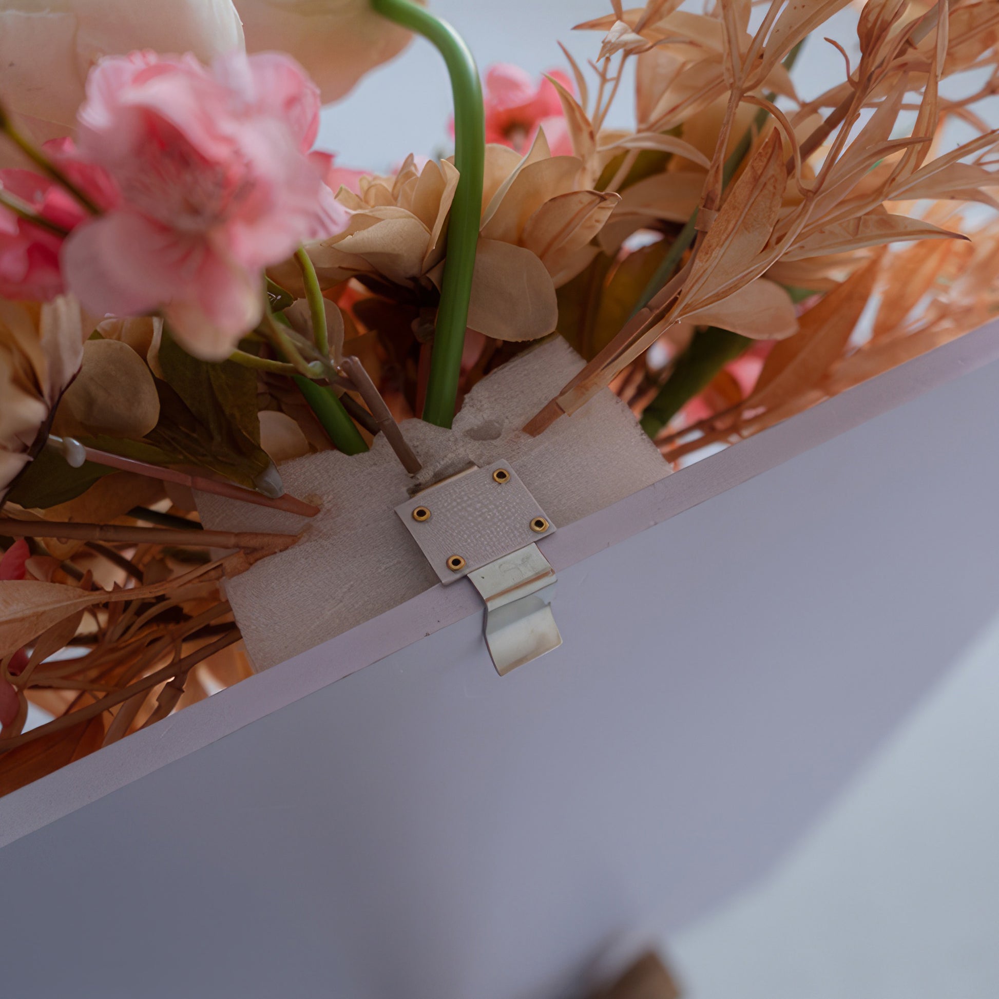 The image displays a close - up of an artificial flower arrangement attached to a board. There's a metal clip with holes, secured by a fabric strip, holding flower stems firmly in place against the board. 