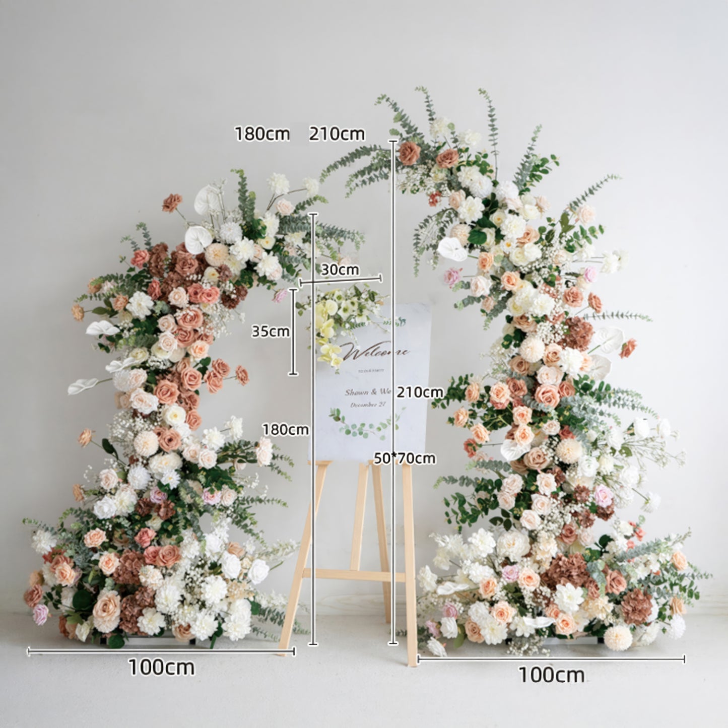 A romantic, asymmetrical floral arch from Valar Flowers, crafted with a soft, warm palette of blush pink, terracotta, and creamy white blooms—including roses, peonies, and delicate baby's breath—intertwined with silvery eucalyptus and lush greenery. A "Welcome" sign on a wooden easel rests between the arch, with clear dimension markings (180cm/210cm height, 100cm base width) for event planning, creating a dreamy, elegant, and sophisticated aesthetic ideal for luxury weddings and editorial photoshoots.