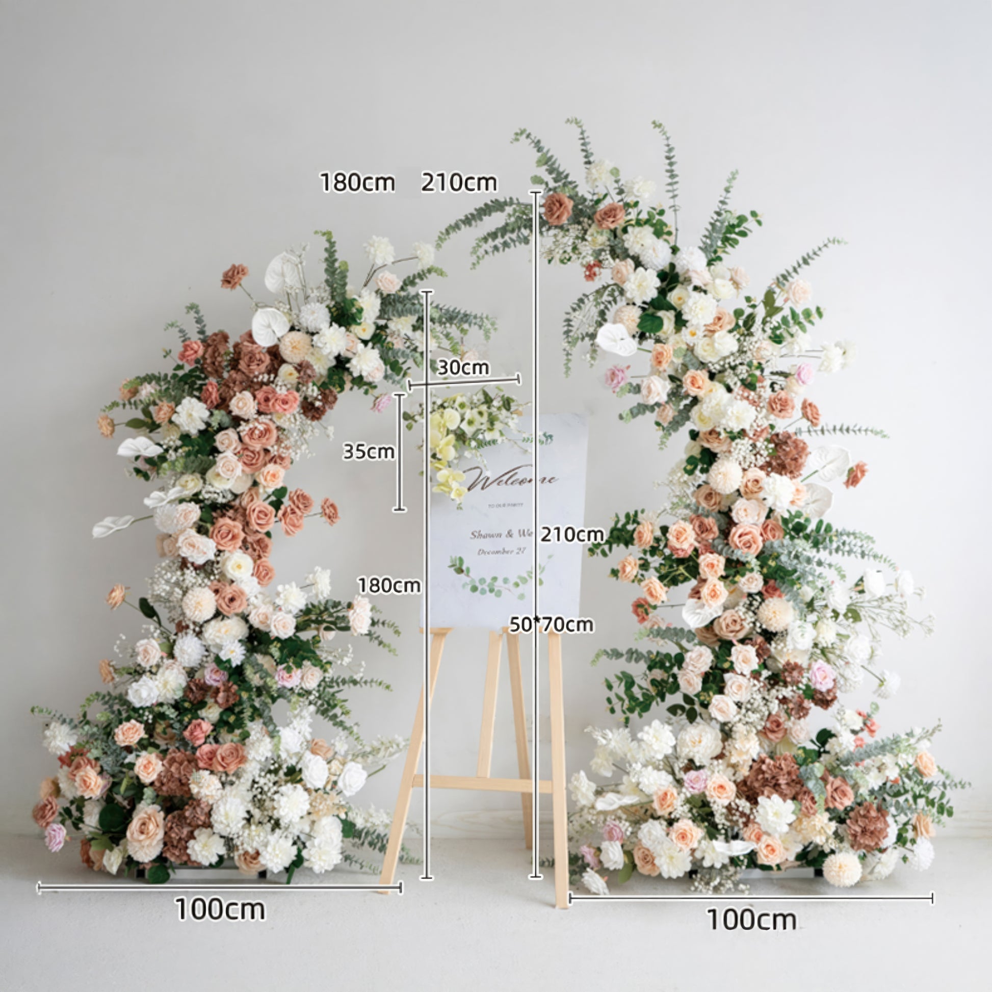 A romantic, asymmetrical floral arch from Valar Flowers, crafted with a soft, warm palette of blush pink, terracotta, and creamy white blooms—including roses, peonies, and delicate baby's breath—intertwined with silvery eucalyptus and lush greenery. A "Welcome" sign on a wooden easel rests between the arch, with clear dimension markings (180cm/210cm height, 100cm base width) for event planning, creating a dreamy, elegant, and sophisticated aesthetic ideal for luxury weddings and editorial photoshoots.