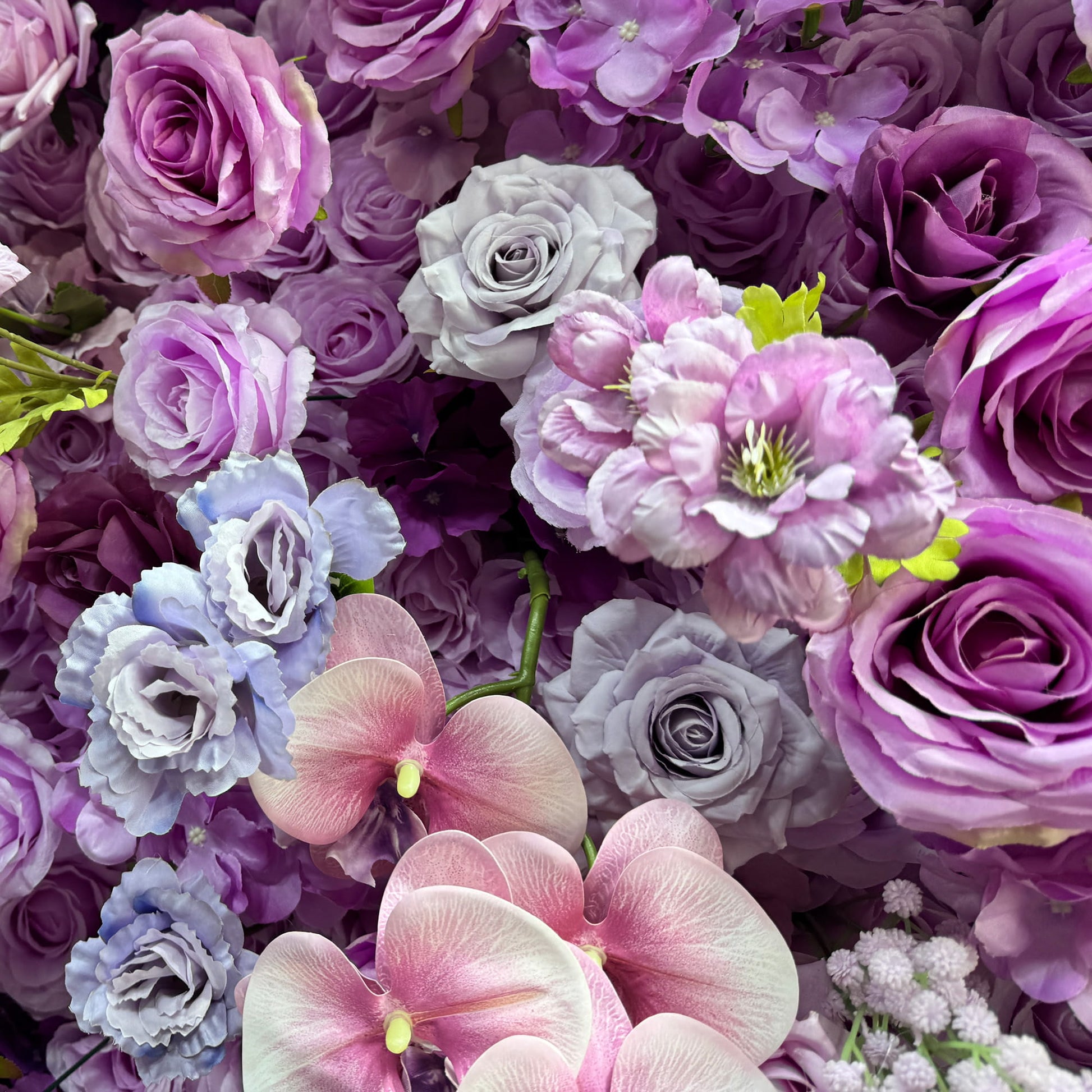 This image showcases a rich tapestry of artificial flowers. Prominent are roses in varying shades of purple, lavender, and deep pink, interspersed with delicate pink - hued orchids and clusters of small, light - colored flowers. The color palette ranges from soft pastels to deeper purples, creating a sumptuous and romantic style, ideal for event backdrops or photo booths. 