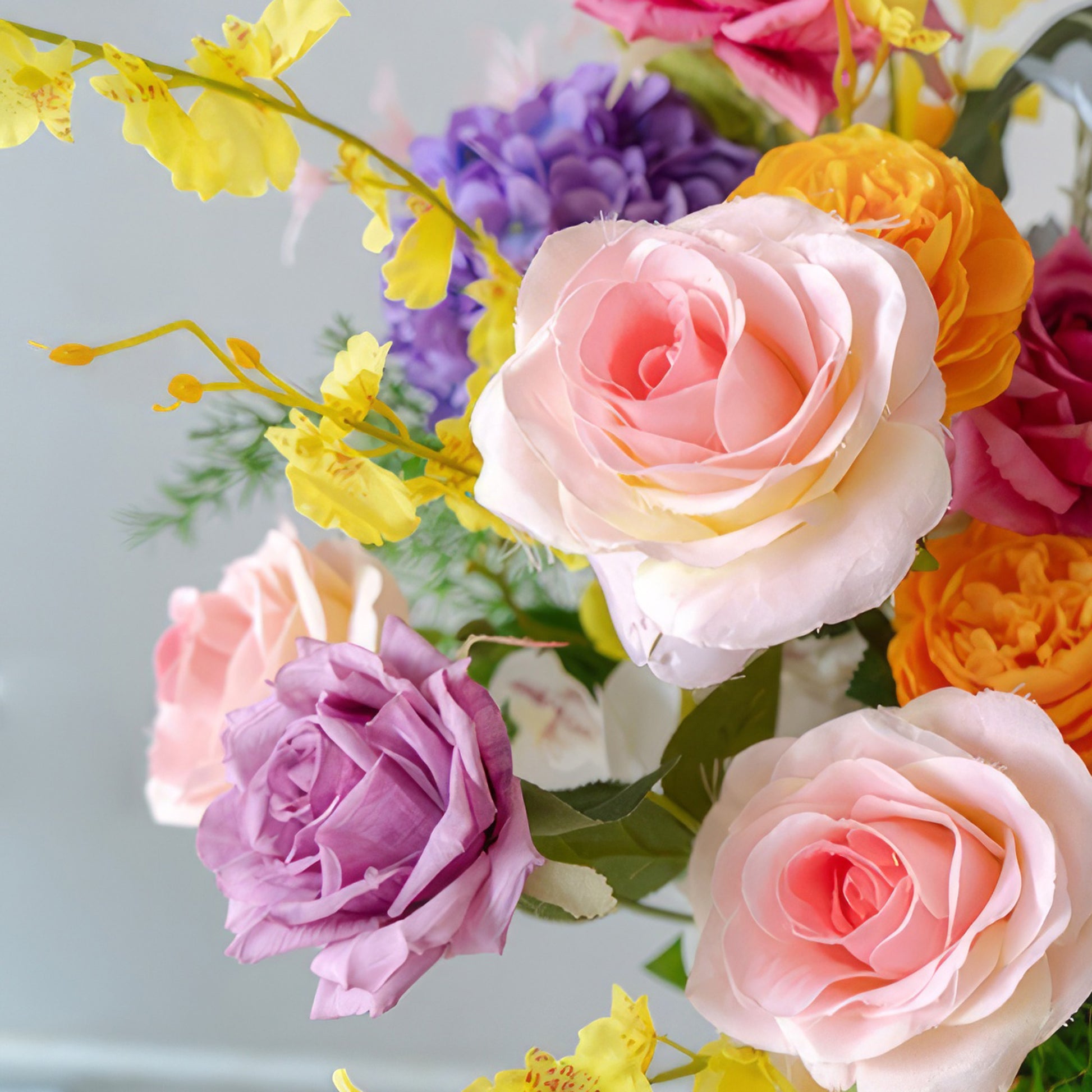 This close - up showcases a beautiful cluster of artificial roses in soft pastel shades like pink and lavender, alongside vivid orange ones. Interspersed are yellow blossoms, adding a pop of bright color. The arrangement exudes a sweet, charming, and romantic style. 