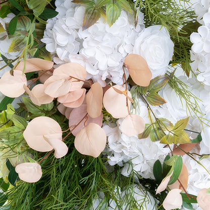 This is a close - up of an artificial flower arrangement. It features pure white hydrangeas and roses, complemented by light peach - colored eucalyptus - like leaves and various green foliage. The style is fresh, elegant, and has a touch of softness. 