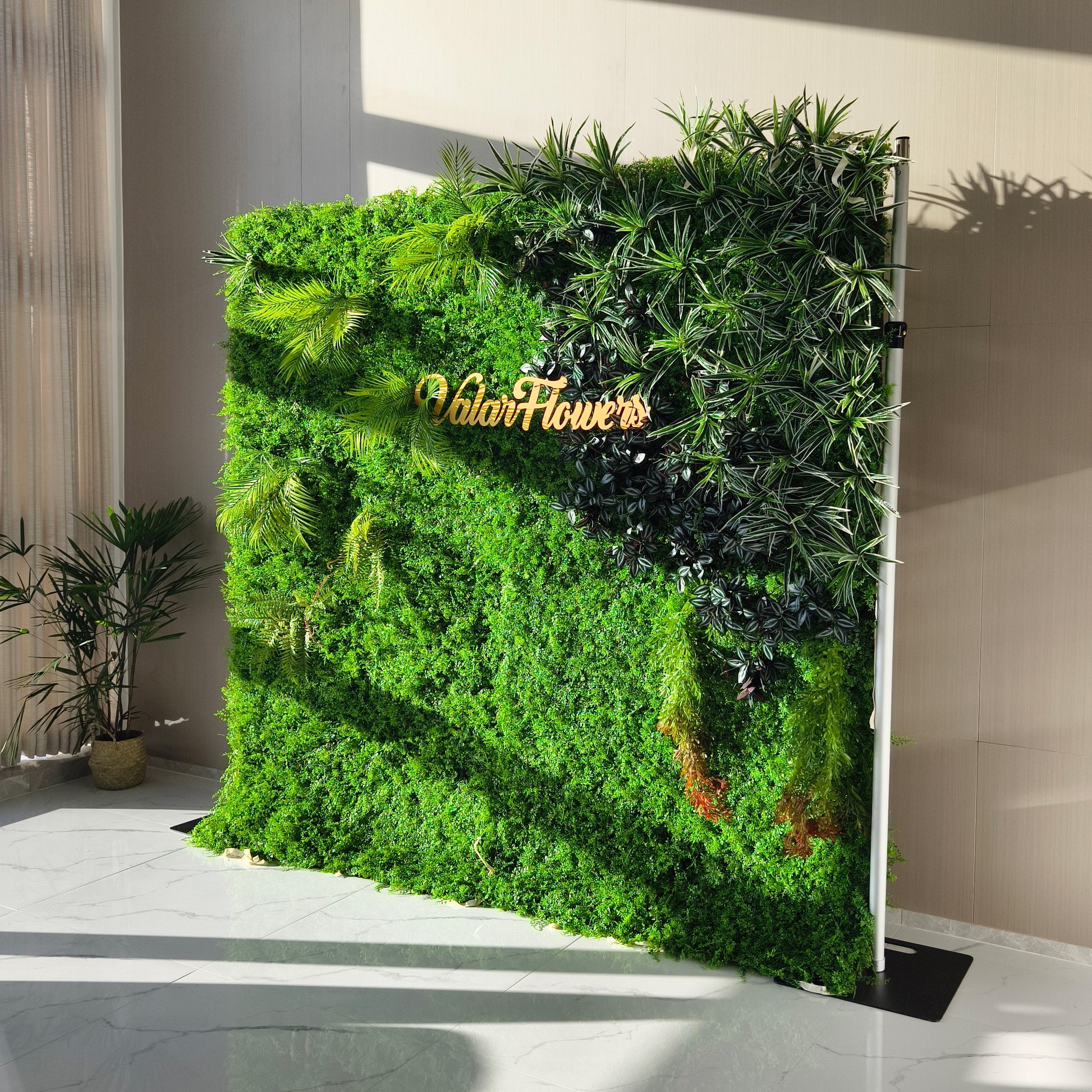 This image showcases a lush artificial greenery wall by ValarFlowers, set indoors on a glossy marble floor. It features a dense base of green foliage, artfully accented with varied plants like palm fronds, textured greens, and variegated leaves. The prominent “ValarFlowers” logo adds a branded touch. With realistic textures and diverse elements, this piece brings organic elegance to interiors, complemented by natural light and a potted plant, exuding a serene and sophisticated atmosphere.