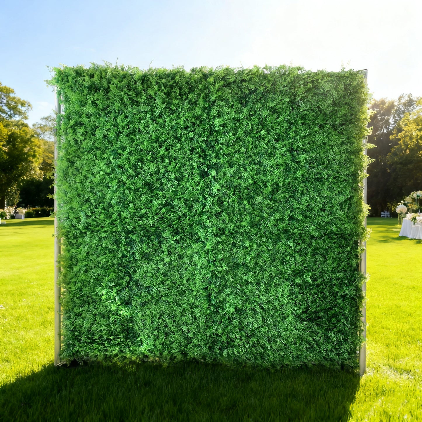 This image showcases a lush artificial greenery backdrop by ValarFlowers, set outdoors on a vibrant lawn. It features dense, uniform green foliage, creating a rich, natural, and visually refreshing display. Bathed in sunlight, with a scenic park and event setup in the background, this piece is perfect for adding organic elegance to outdoor events, exuding a serene and sophisticated atmosphere.
