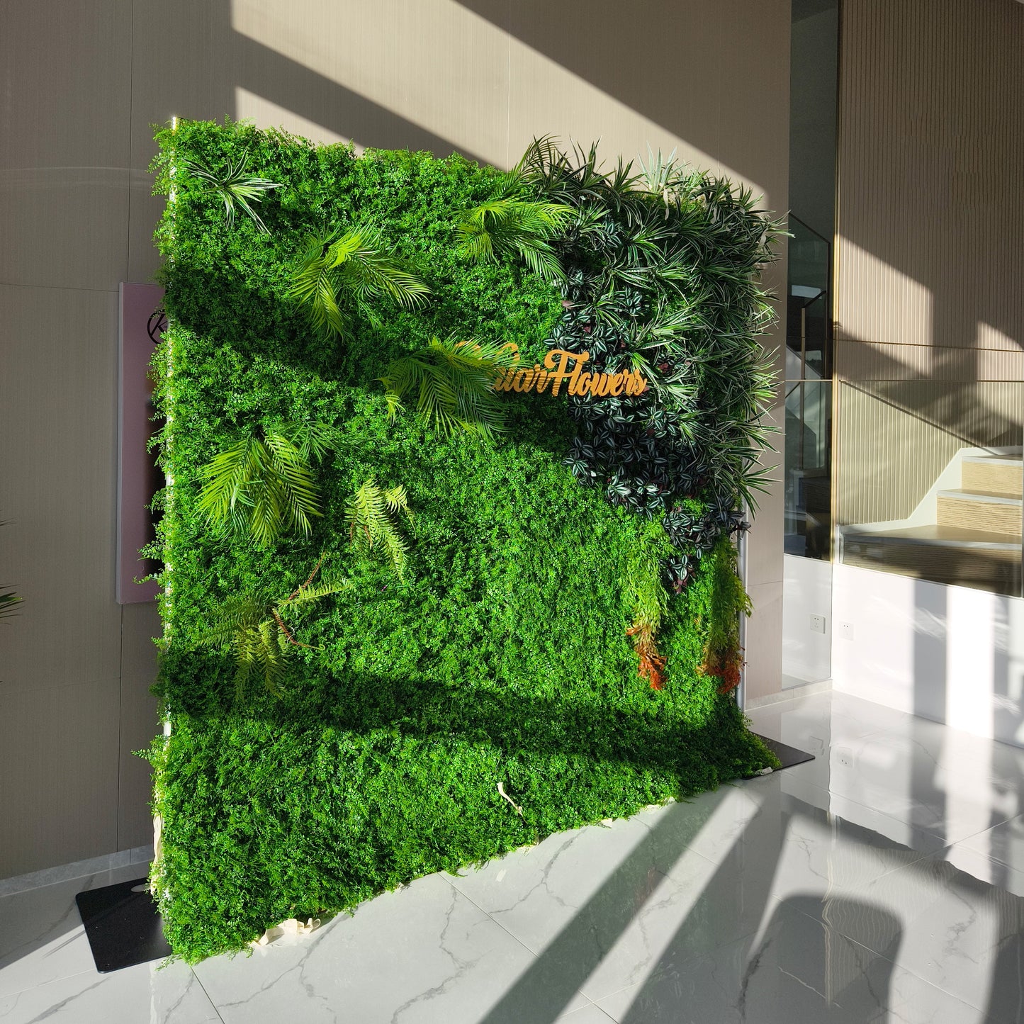 This image showcases a lush artificial greenery wall by ValarFlowers, set indoors on a glossy marble floor. It features a dense base of green foliage, artfully accented with varied plants like palm fronds, ferns, and textured greens. The prominent “ValarFlowers” logo adds a branded touch. With realistic textures and diverse elements, this piece brings organic elegance to the space, complemented by natural light and modern architecture, exuding a serene and sophisticated atmosphere.