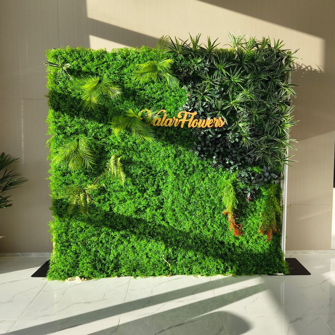This image showcases a lush artificial greenery wall by ValarFlowers, set indoors on a glossy marble floor. It features a dense base of green foliage, artfully accented with varied plants like palm fronds, ferns, and textured greens. The prominent “ValarFlowers” logo adds a branded touch. With realistic textures and diverse elements, this piece brings organic elegance to interiors, exuding a serene and sophisticated atmosphere.