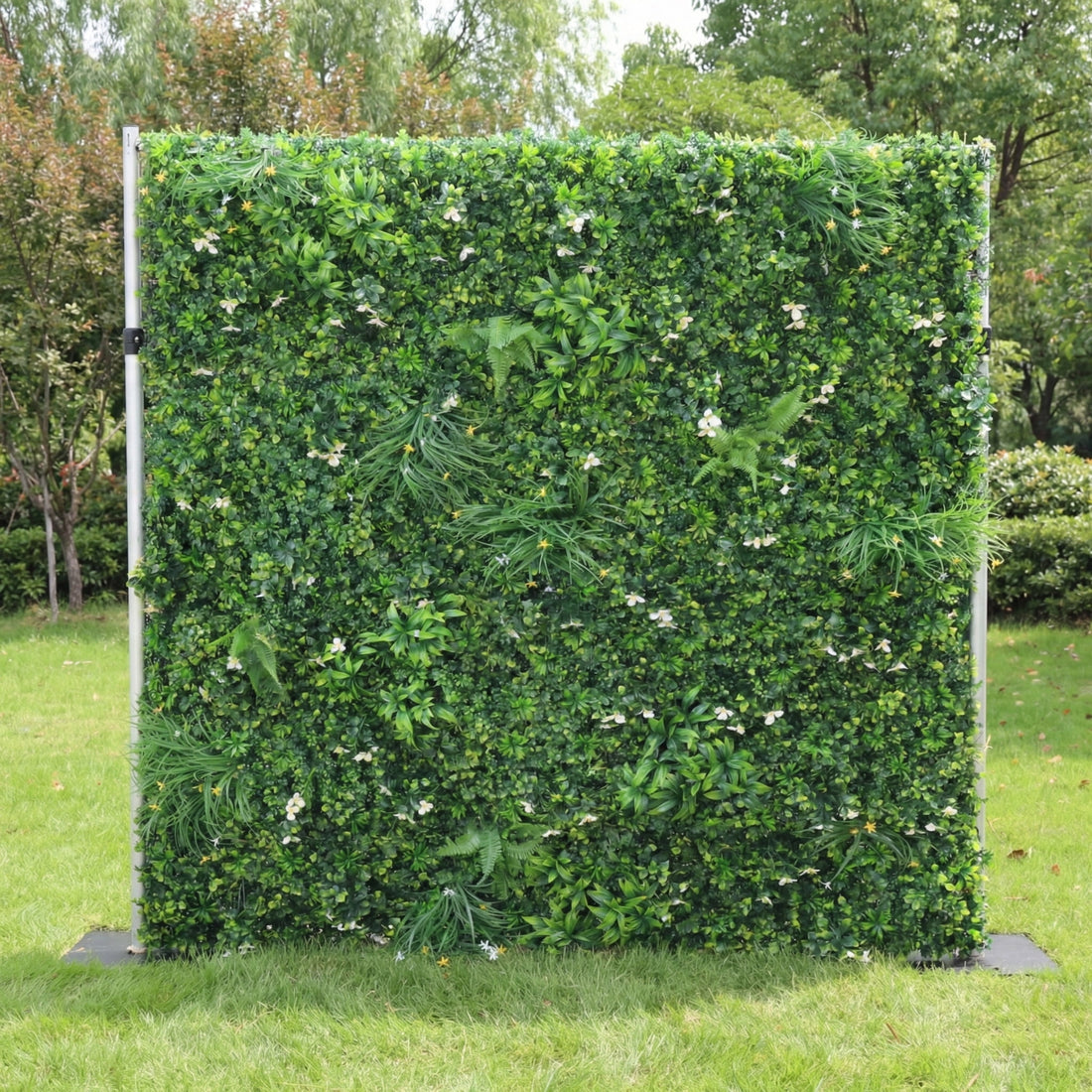 This outdoor greenery wall from Valar Flowers is a lush, textured backdrop of dense, vibrant green foliage—including ferns, leafy shrubs, and grassy accents—interspersed with delicate white and yellow wildflowers. Set on a sunlit green lawn framed by leafy trees, the design evokes a fresh, natural, and elegantly understated atmosphere ideal for weddings, photo backdrops, or elegant outdoor celebrations.
