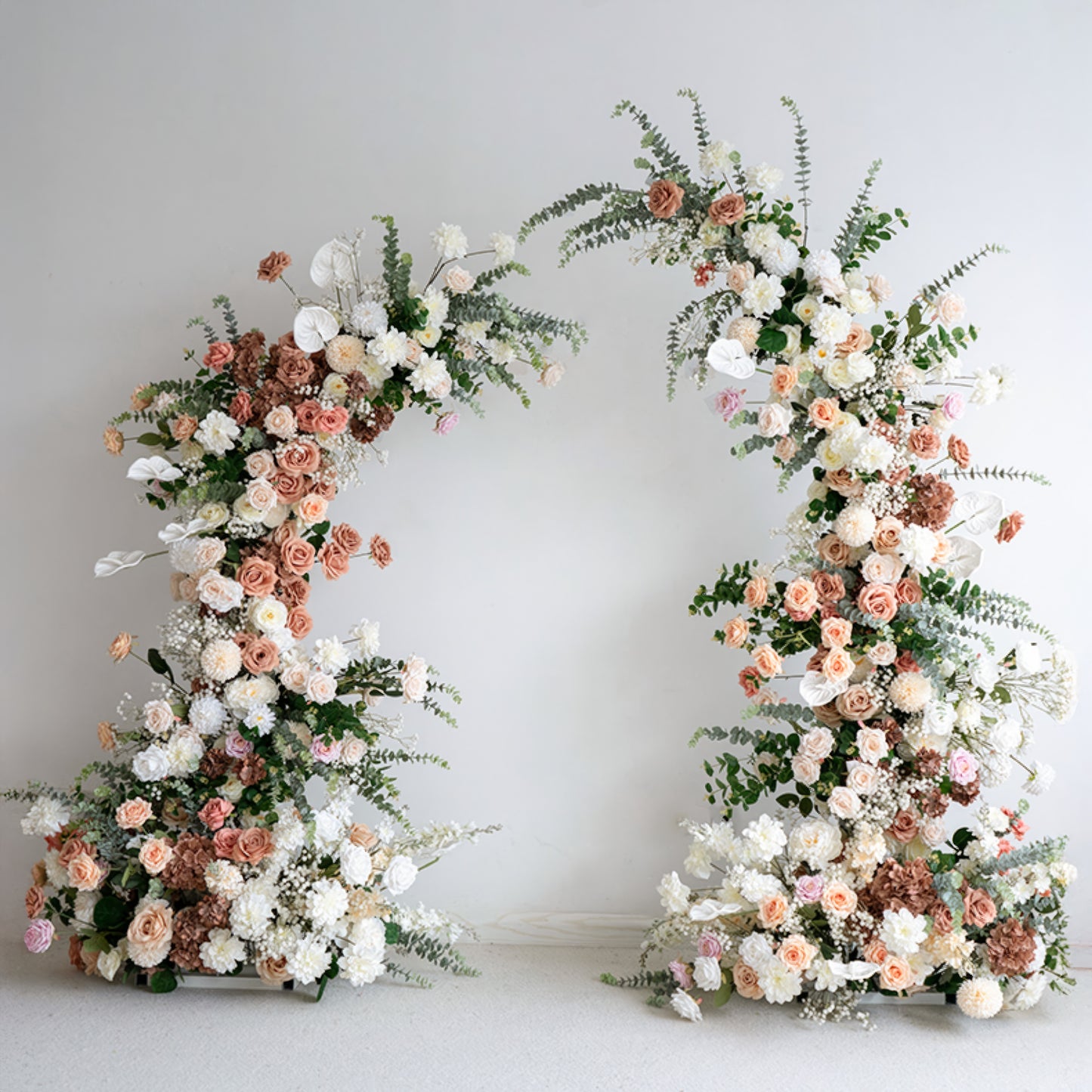 A romantic, asymmetrical floral arch from Valar Flowers, crafted with a soft, warm palette of blush pink, terracotta, and creamy white blooms—including roses, peonies, and delicate baby's breath—intertwined with silvery eucalyptus and lush greenery. Set against a neutral studio backdrop, it creates a dreamy, elegant, and sophisticated aesthetic ideal for luxury weddings and editorial photoshoots.
