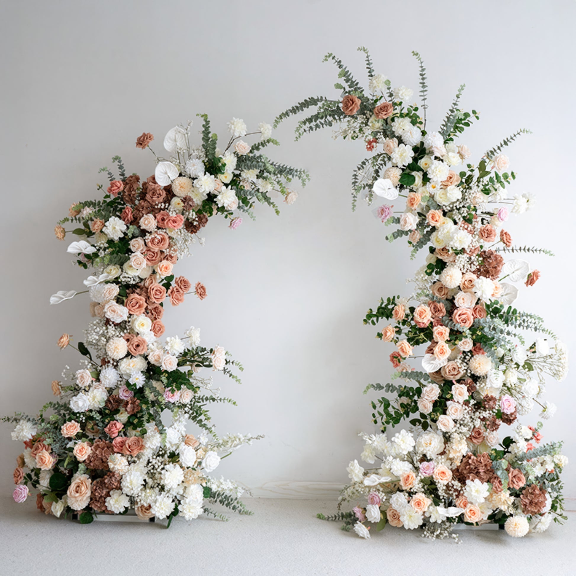 A romantic, asymmetrical floral arch from Valar Flowers, crafted with a soft, warm palette of blush pink, terracotta, and creamy white blooms—including roses, peonies, and delicate baby's breath—intertwined with silvery eucalyptus and lush greenery. Set against a neutral studio backdrop, it creates a dreamy, elegant, and sophisticated aesthetic ideal for luxury weddings and editorial photoshoots.
