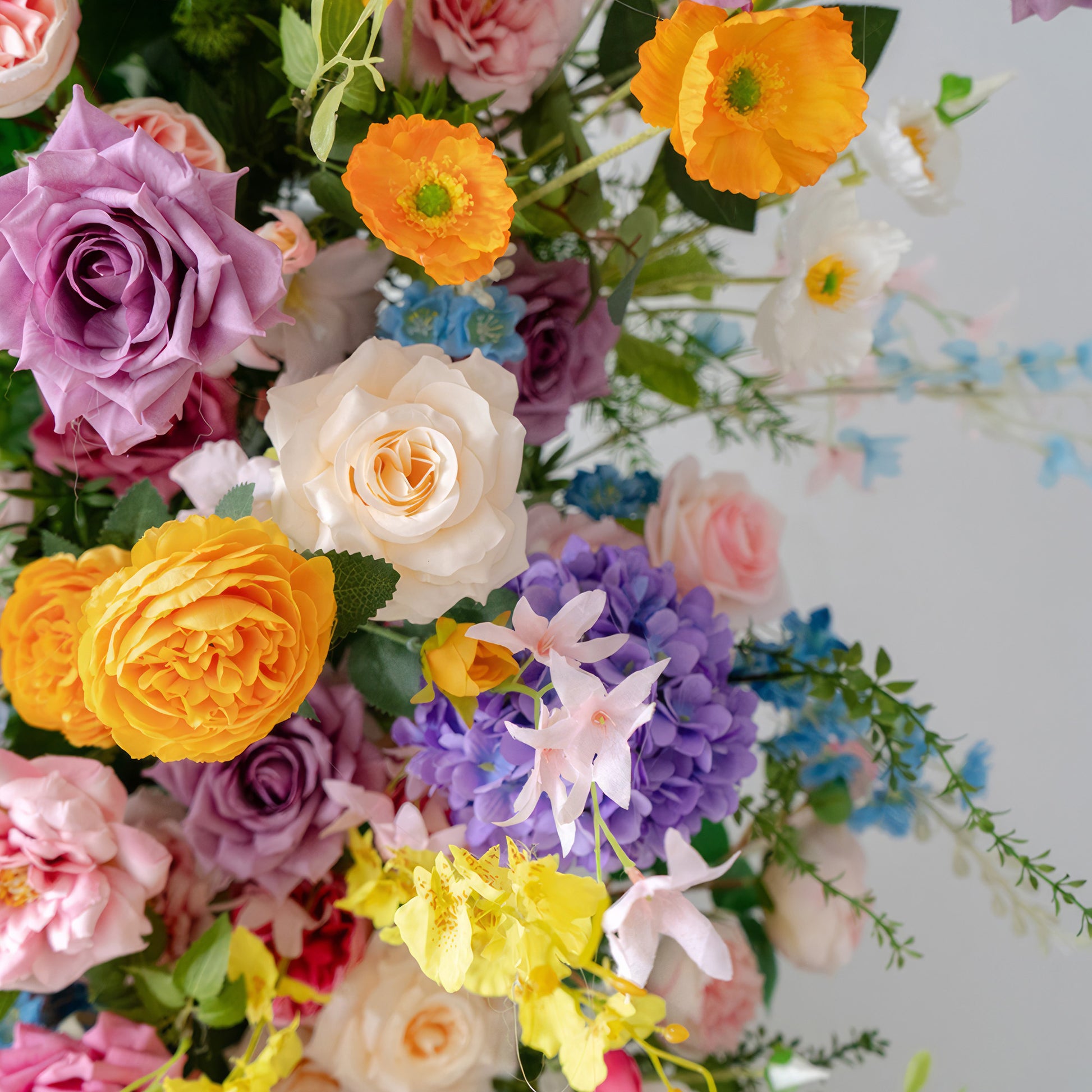 This image features a vibrant mix of artificial flowers. There are roses in hues of purple, pink, and yellow, along with bright orange poppies and purple hydrangeas. Small blue and yellow blooms add to the variety, creating a lively and colorful arrangement with a cheerful, festive style. 