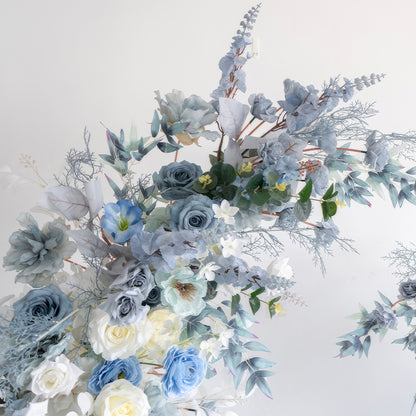 This image features an artificial flower arrangement with a cool and calming color scheme of various blues and whites. It includes blue roses, white roses, and other blue - toned blooms, complemented by different types of foliage. The overall style is elegant, serene, and gives a sense of tranquility. 