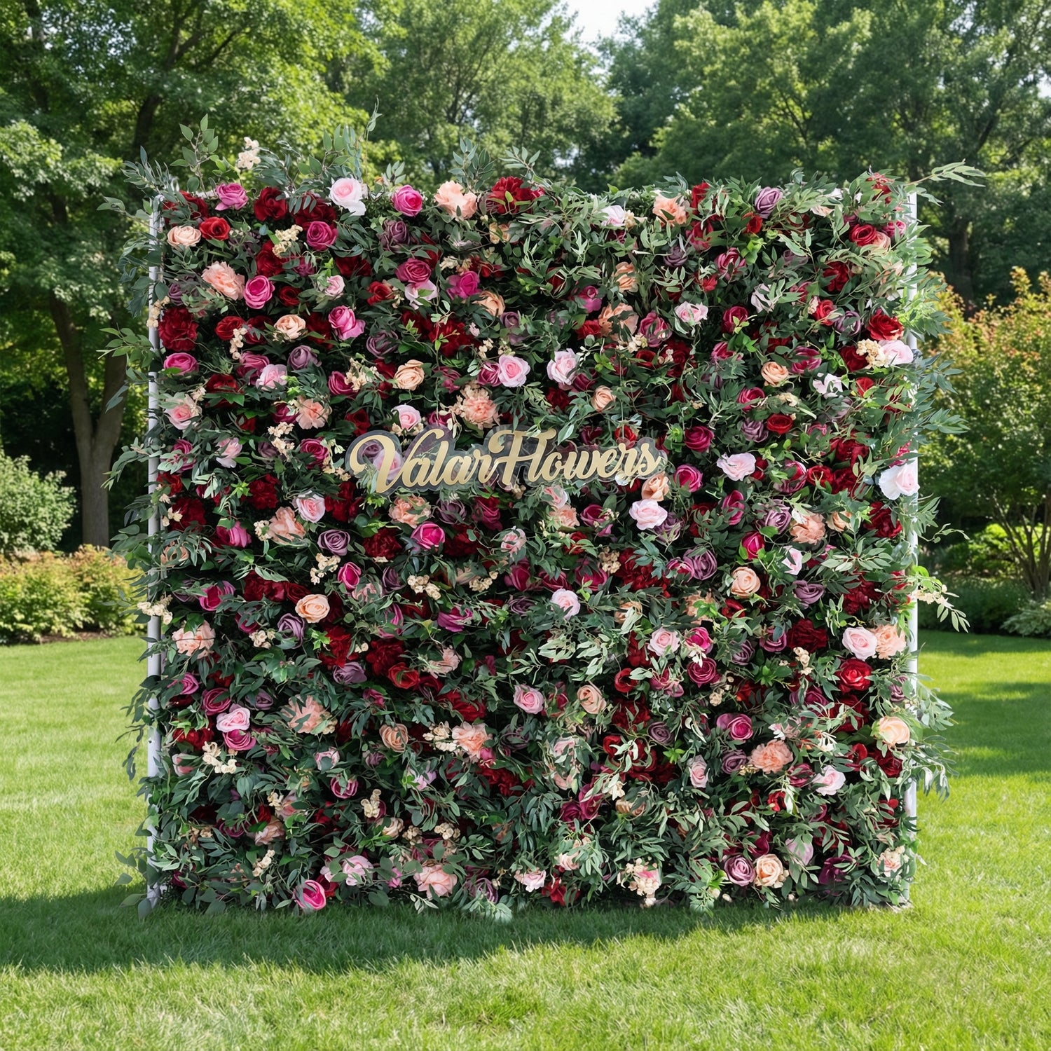 This image showcases a vibrant flower wall by Valar Flowers, featuring a dense arrangement of artificial roses in rich reds, pinks, and peaches, complemented by green foliage. Set outdoors on lush grass with trees in the background, a gold “Valar Flowers” sign is integrated at the center. It exudes elegance and sophistication, ideal for events, highlighting the brand’s craftsmanship in creating eye-catching floral decor.
