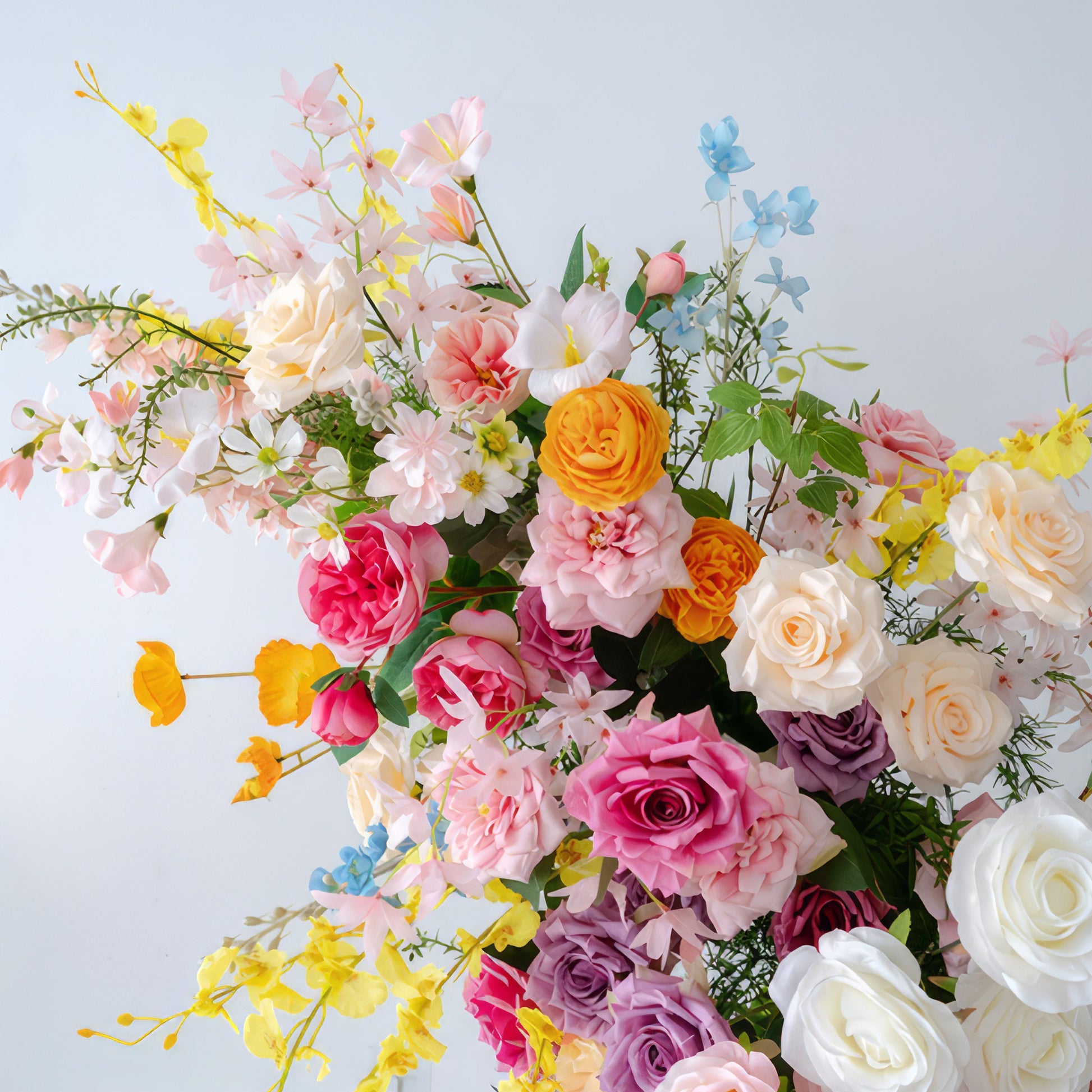 This floral arrangement is a vivid symphony of colors. It combines roses in various shades like white, pink, purple, and orange, along with delicate flowers in soft yellow, blue, and pale pink. Green foliage enhances the natural look, presenting a lively, cheerful, and celebratory style. 