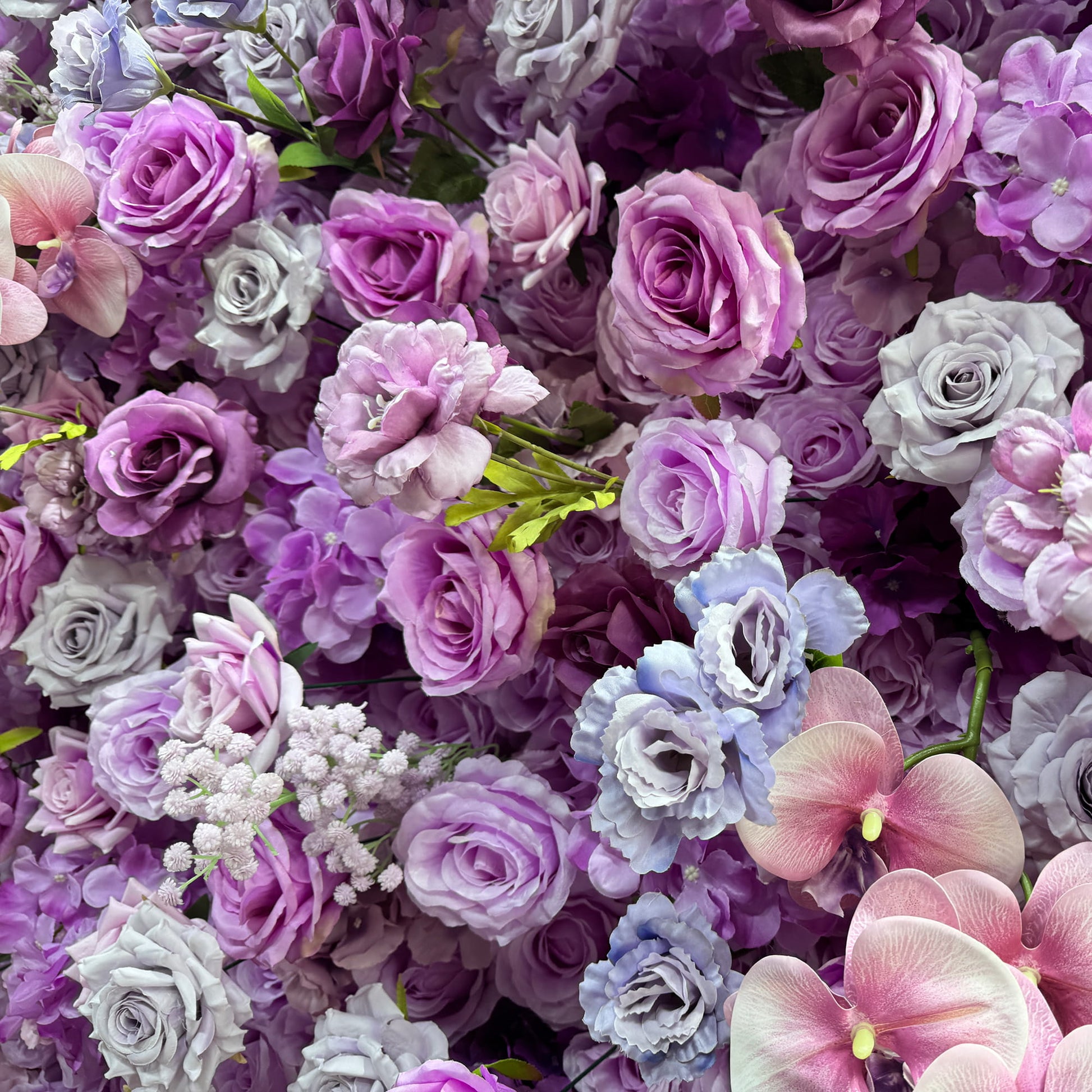 This image features a dense arrangement of artificial flowers. Prominent among them are roses in various shades of purple, ranging from deep violet to light lavender, along with some pink - tinged orchids and clusters of small white flowers resembling baby's breath. The color palette is a harmonious blend of cool purple tones, creating a romantic and elegant style, perfect for event decorations. 
