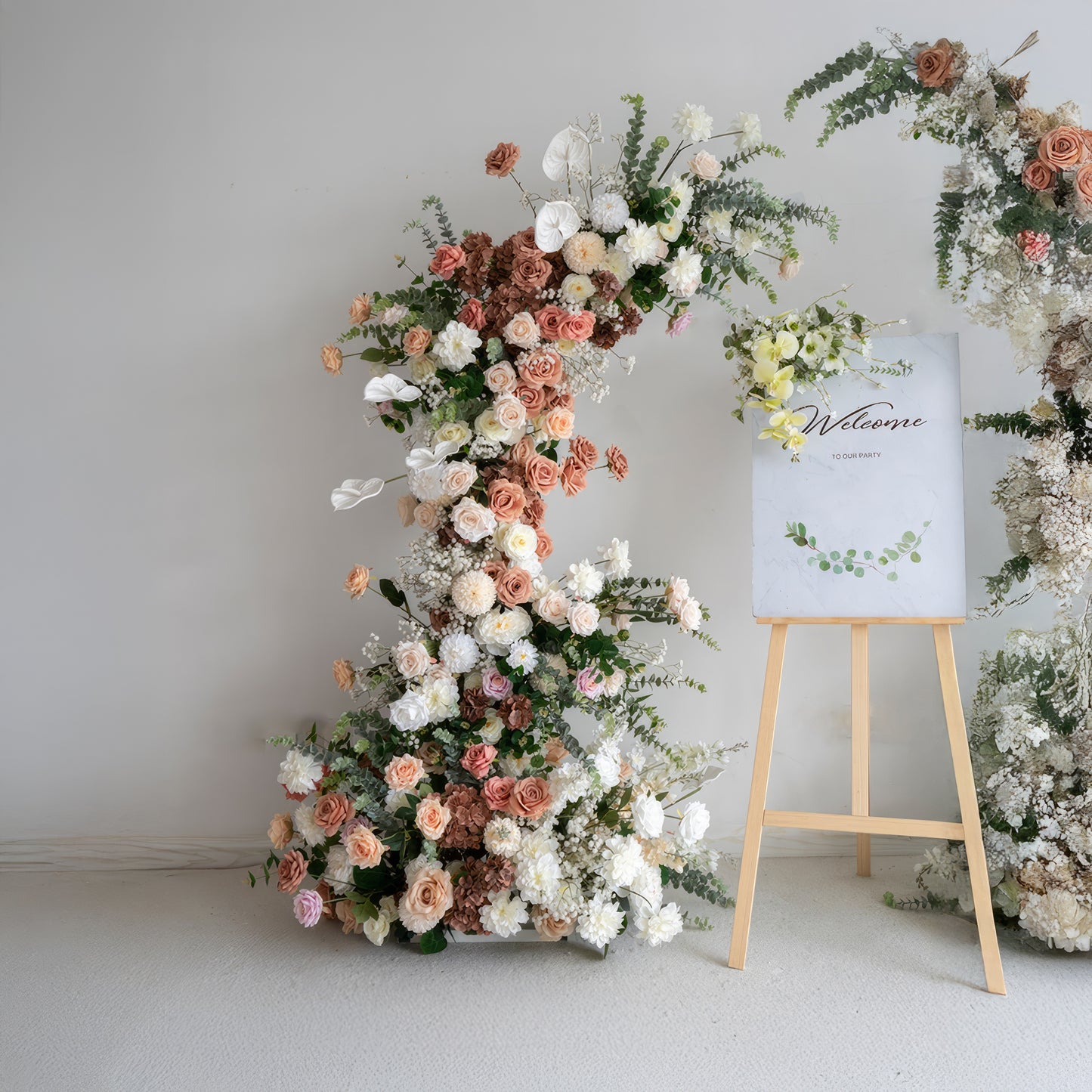 A romantic, asymmetrical floral arch from Valar Flowers, crafted with a soft, warm palette of blush pink, terracotta, and creamy white blooms—including roses, peonies, and delicate baby's breath—intertwined with silvery eucalyptus and lush greenery. A "Welcome" sign on a wooden easel rests beside the arch, creating a dreamy, elegant, and sophisticated aesthetic ideal for luxury weddings and editorial photoshoots.
