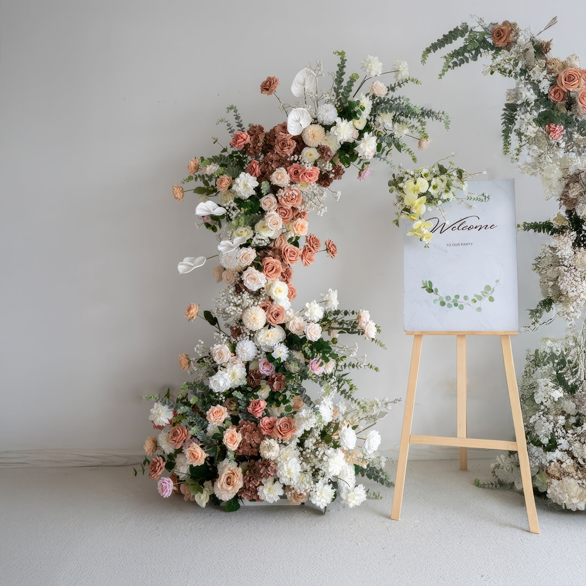 A romantic, asymmetrical floral arch from Valar Flowers, crafted with a soft, warm palette of blush pink, terracotta, and creamy white blooms—including roses, peonies, and delicate baby's breath—intertwined with silvery eucalyptus and lush greenery. A "Welcome" sign on a wooden easel rests beside the arch, creating a dreamy, elegant, and sophisticated aesthetic ideal for luxury weddings and editorial photoshoots.
