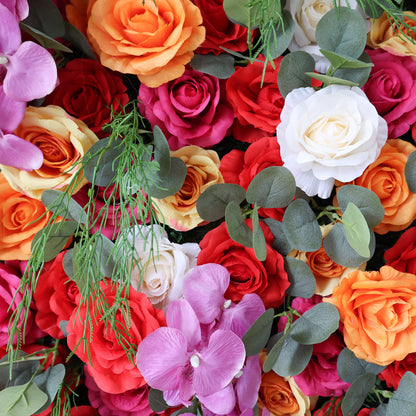This image showcases a vibrant floral arrangement by Valar Flowers. It features a dense cluster of artificial flowers in a rich palette of red, orange, pink, and white, including roses and orchids, interspersed with lush green foliage. The blooms are intricately arranged to create a lush, textured, and visually striking display. Perfect for adding a touch of energy and elegance to events or spaces, the arrangement exudes a lively and sophisticated atmosphere.