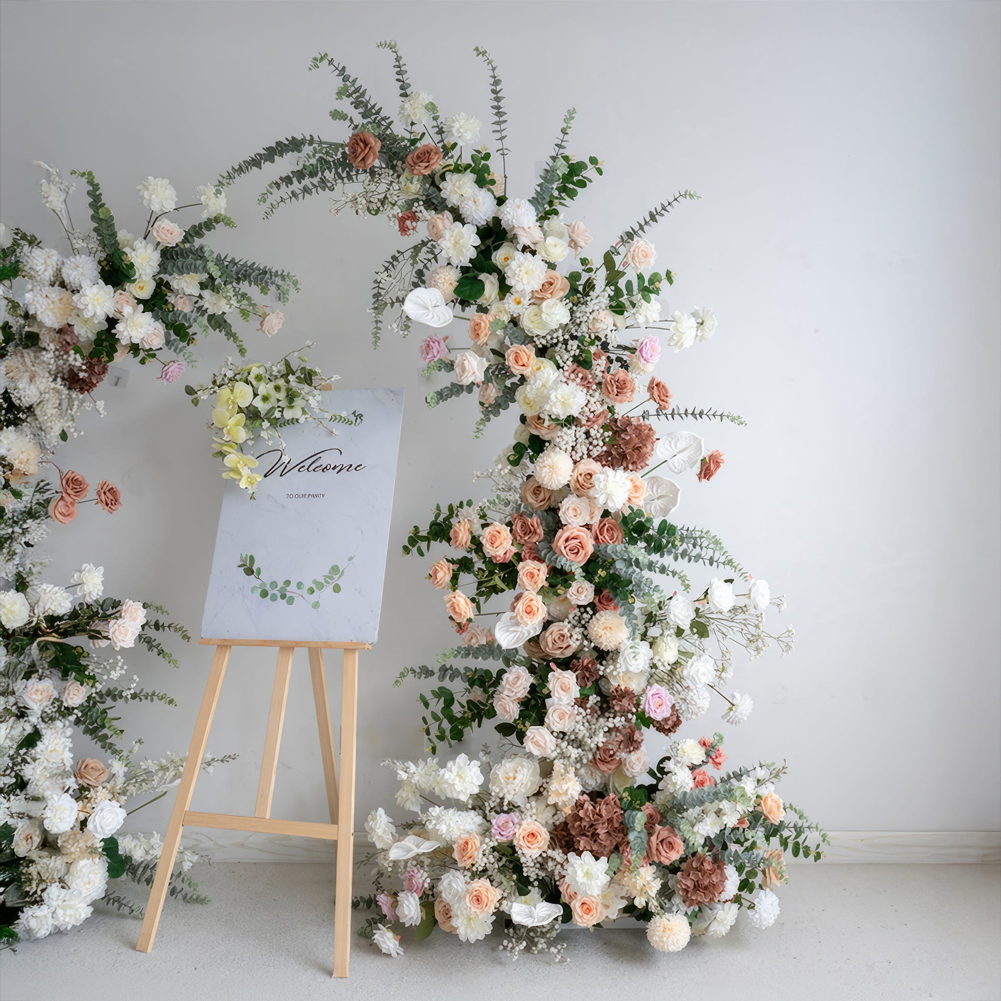 A romantic, asymmetrical floral arch from Valar Flowers, crafted with a soft, warm palette of blush pink, terracotta, and creamy white blooms—including roses, peonies, and delicate baby's breath—intertwined with silvery eucalyptus and lush greenery. A "Welcome" sign on a wooden easel rests beside the arch, creating a dreamy, elegant, and sophisticated aesthetic ideal for luxury weddings and editorial photoshoots.
